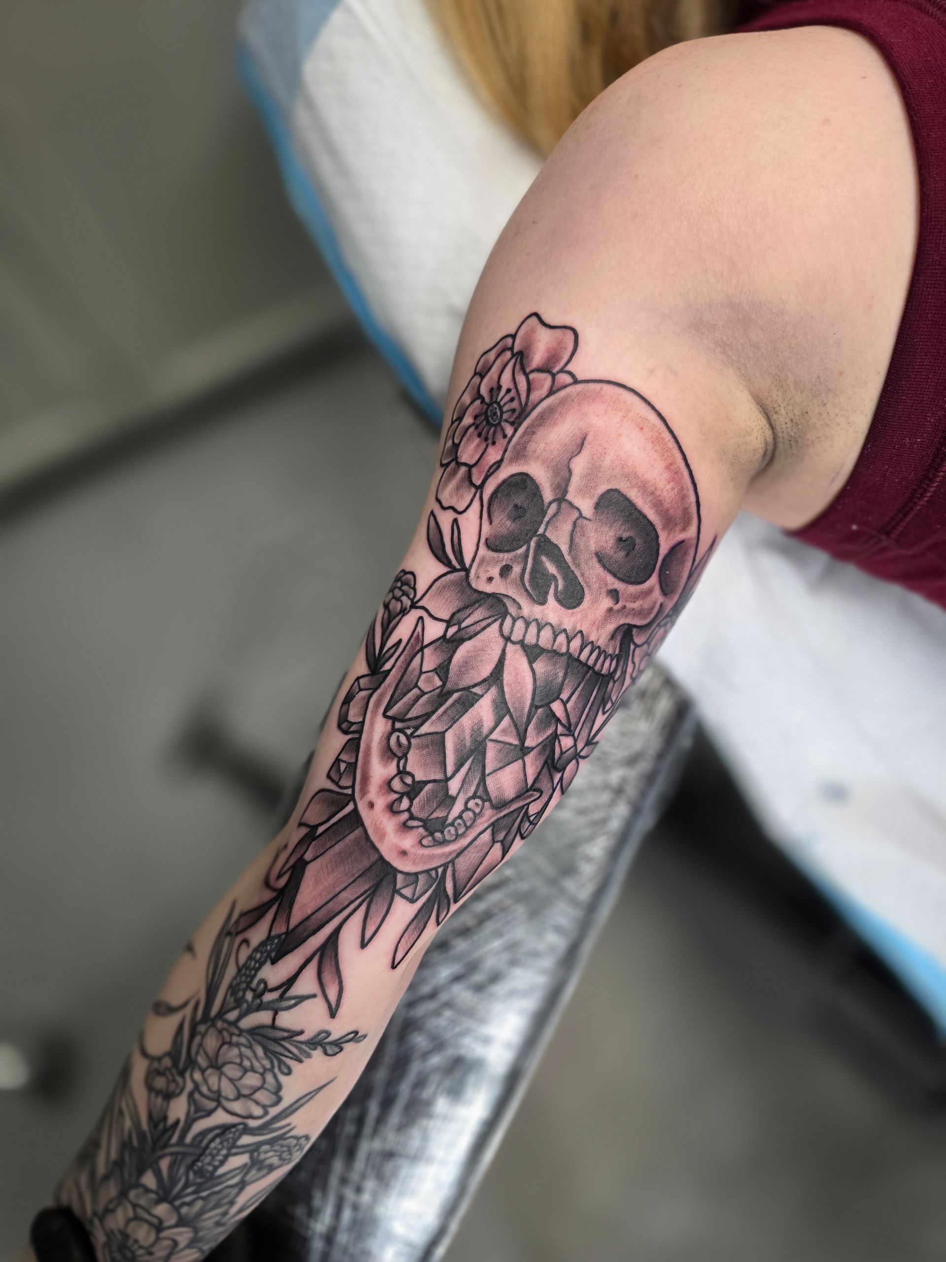 Skull tattoo on a person's forearm and upper arm with floral elements; black and gray ink.