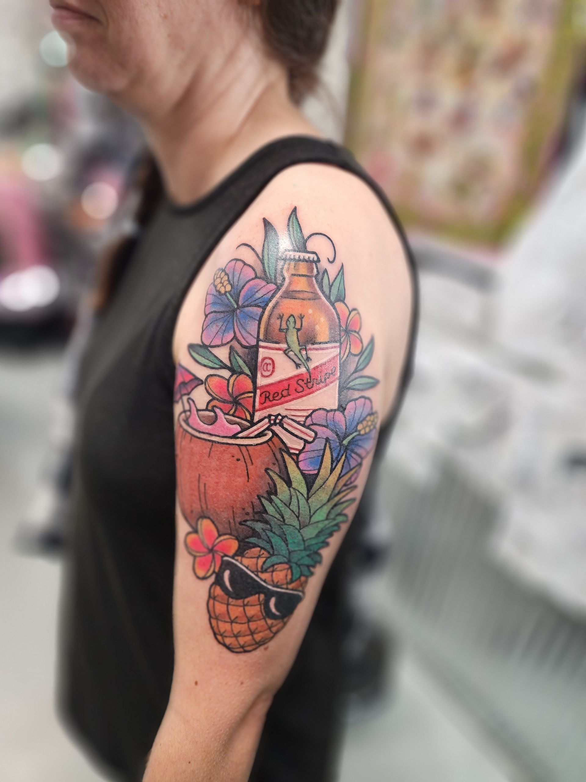 Tattoo of a beer bottle, coconut, and sunglasses-wearing pineapple with flowers on a person's arm.