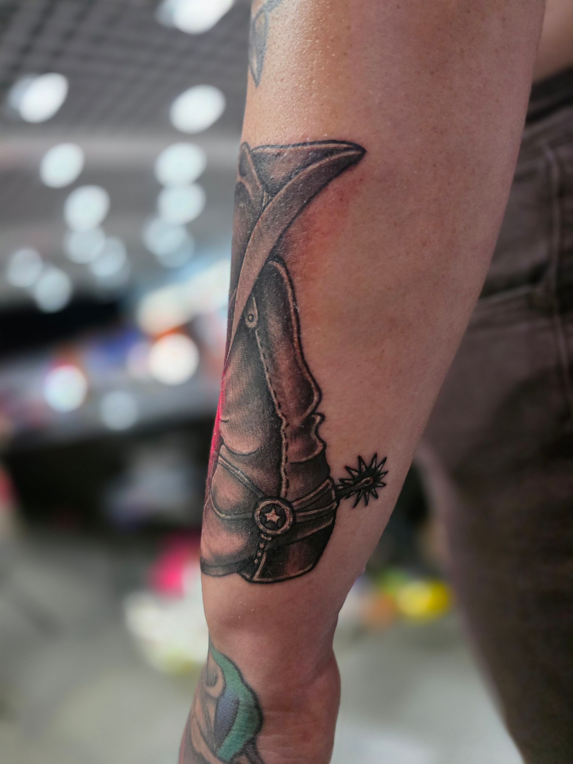 Arm with a black and gray cowboy boot tattoo featuring a spur.
