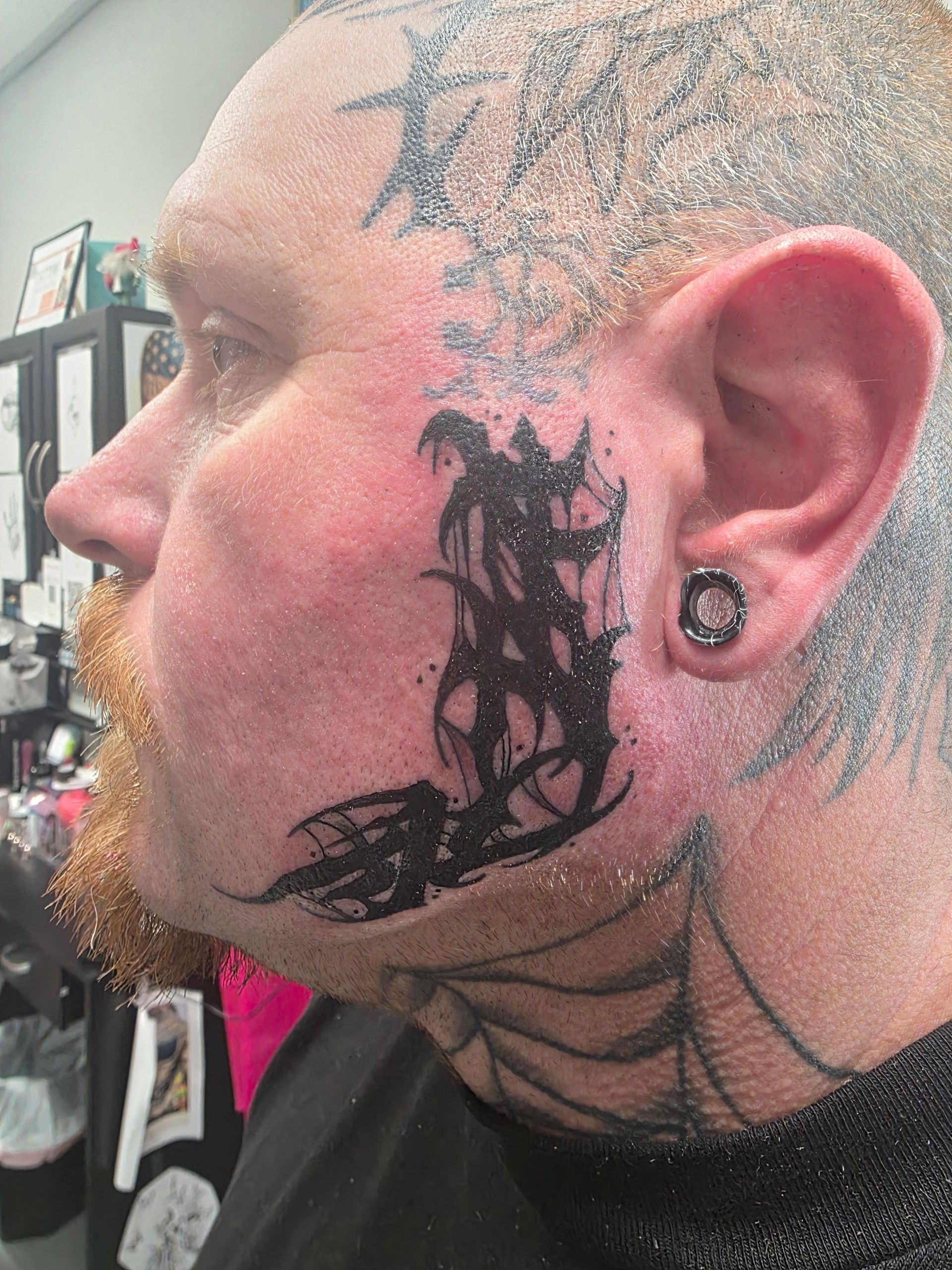 Man with face tattoo; black ink design on cheek, ear gauge, and hairline tattoo.