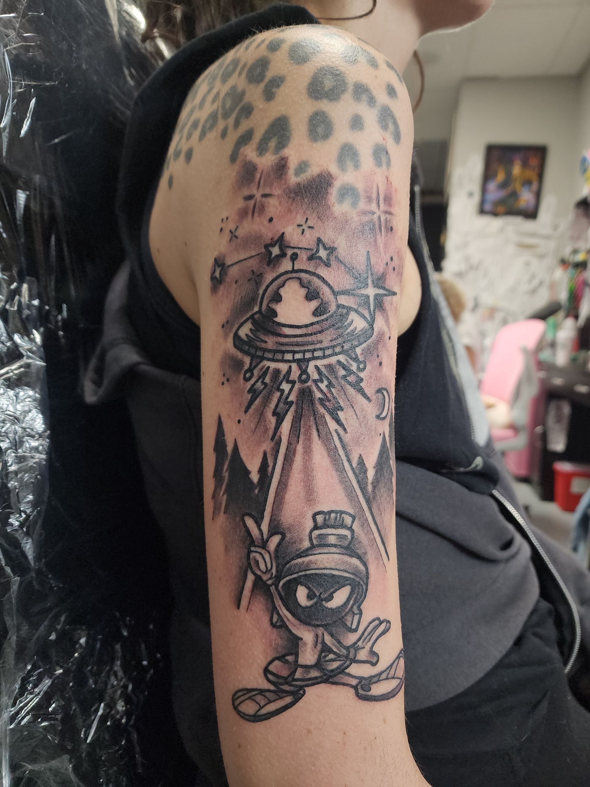 Tattoo of Marvin the Martian with UFO beaming down on a forest scene. Tattoo extends onto the arm with a leopard print design.