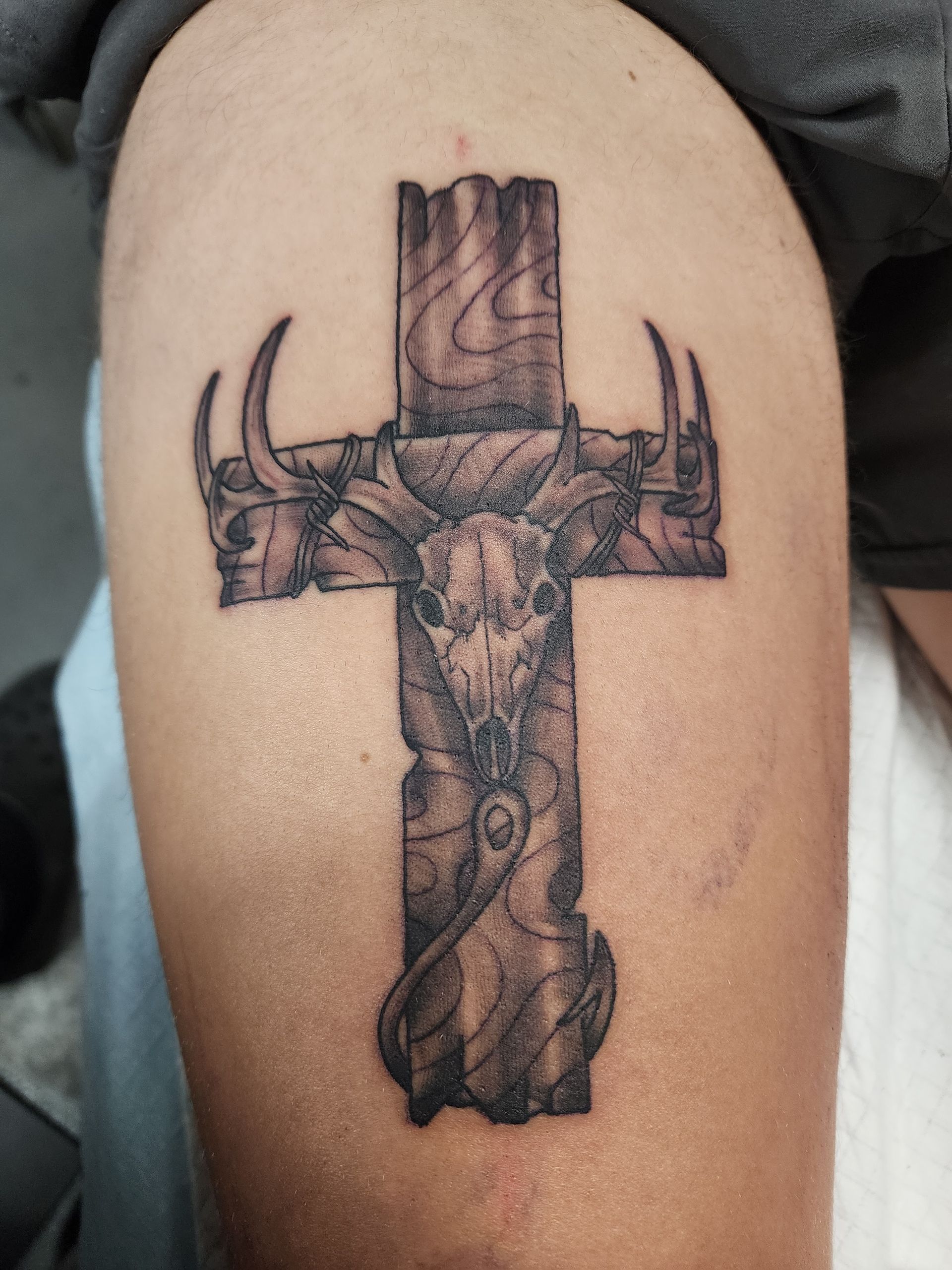 Tattoo on an arm: a textured cross with animal skull and horns.