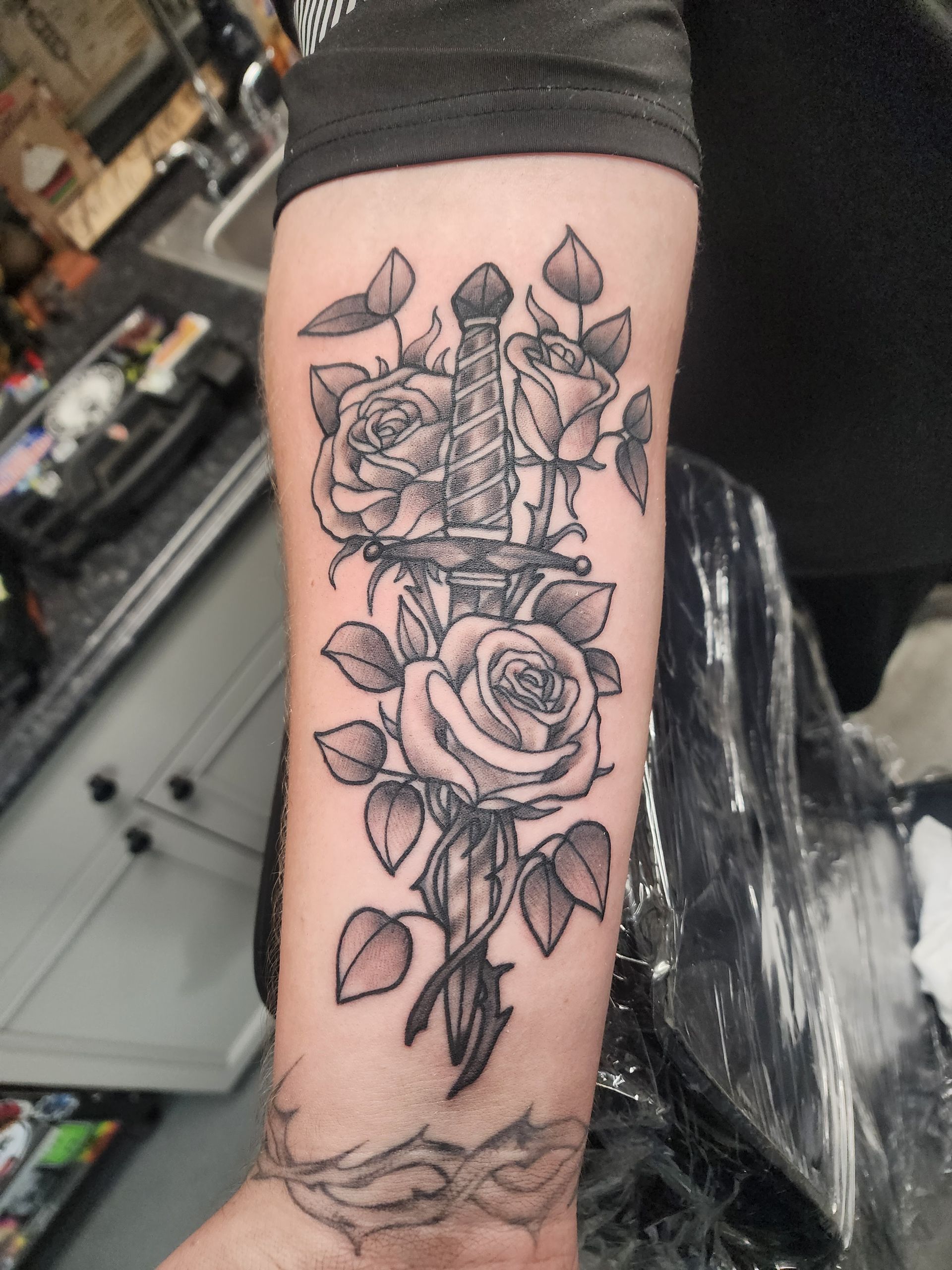 Tattoo of a dagger piercing roses on a forearm, black and white ink, set in a tattoo studio.
