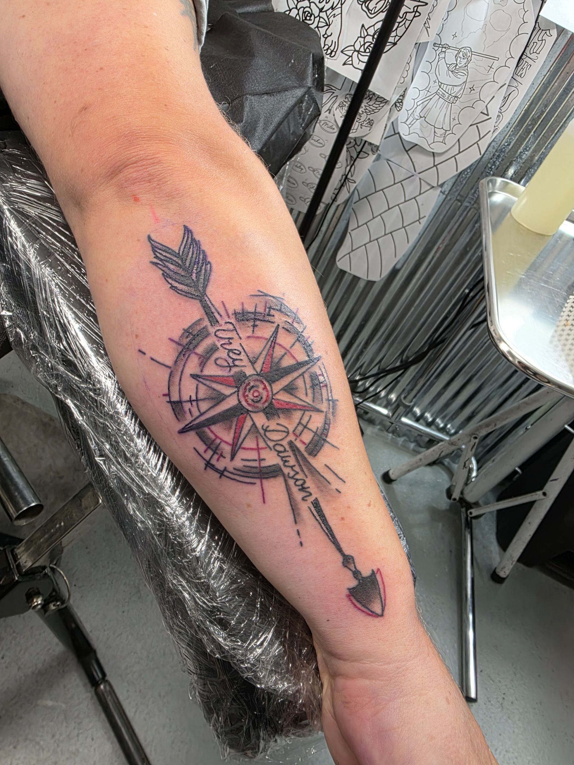 Tattoo of a compass with an arrow on a person's forearm. Black ink with red accents, in a studio setting.