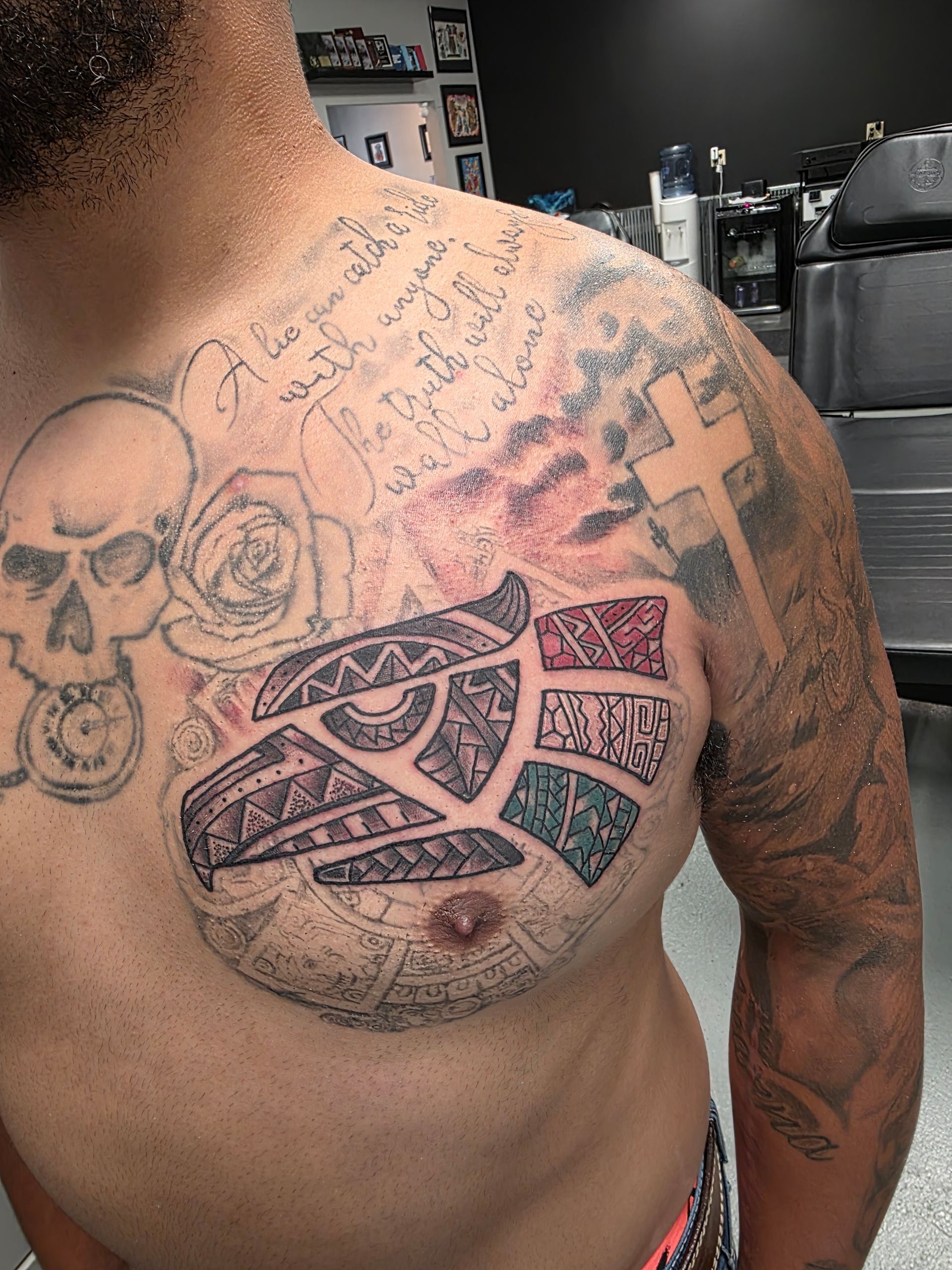 Man's chest tattoo with a geometric bird design, skull, roses, cross, and script against a blurred background.