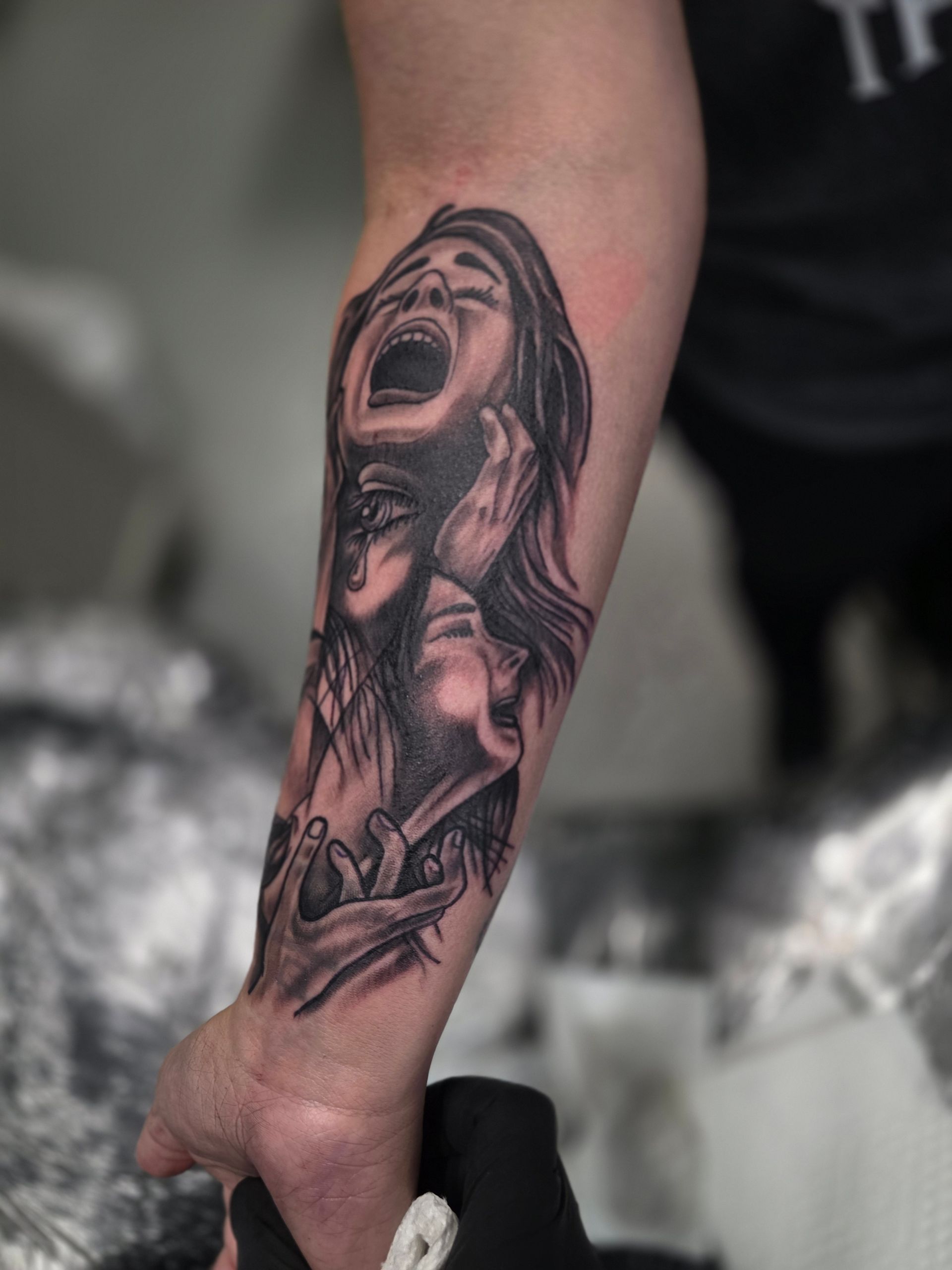 Black and white tattoo of faces expressing anguish on a forearm, with hands and arm holding the arm.