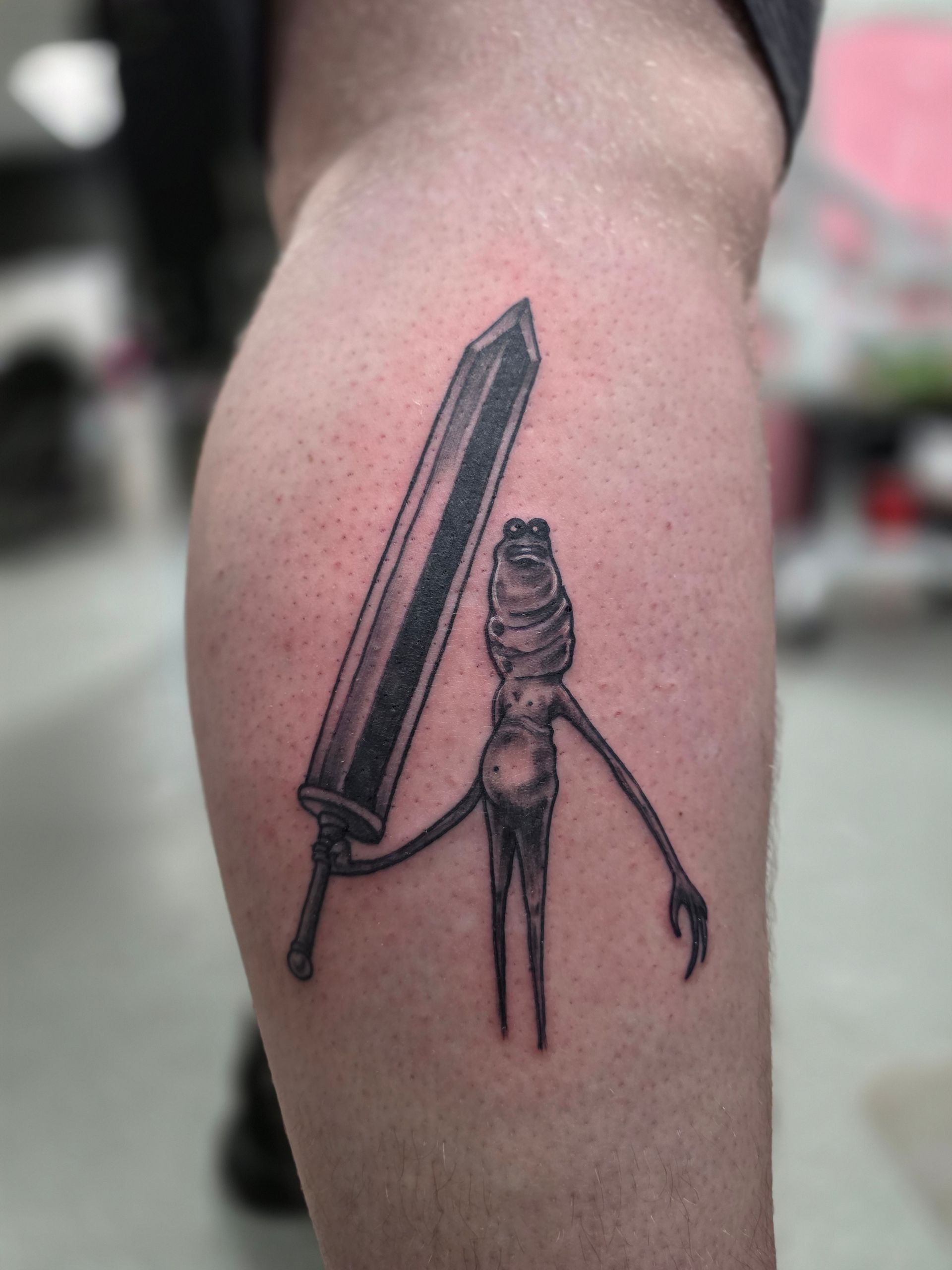 Tattoo of a thin figure holding a large sword on a person's calf. Black and gray.
