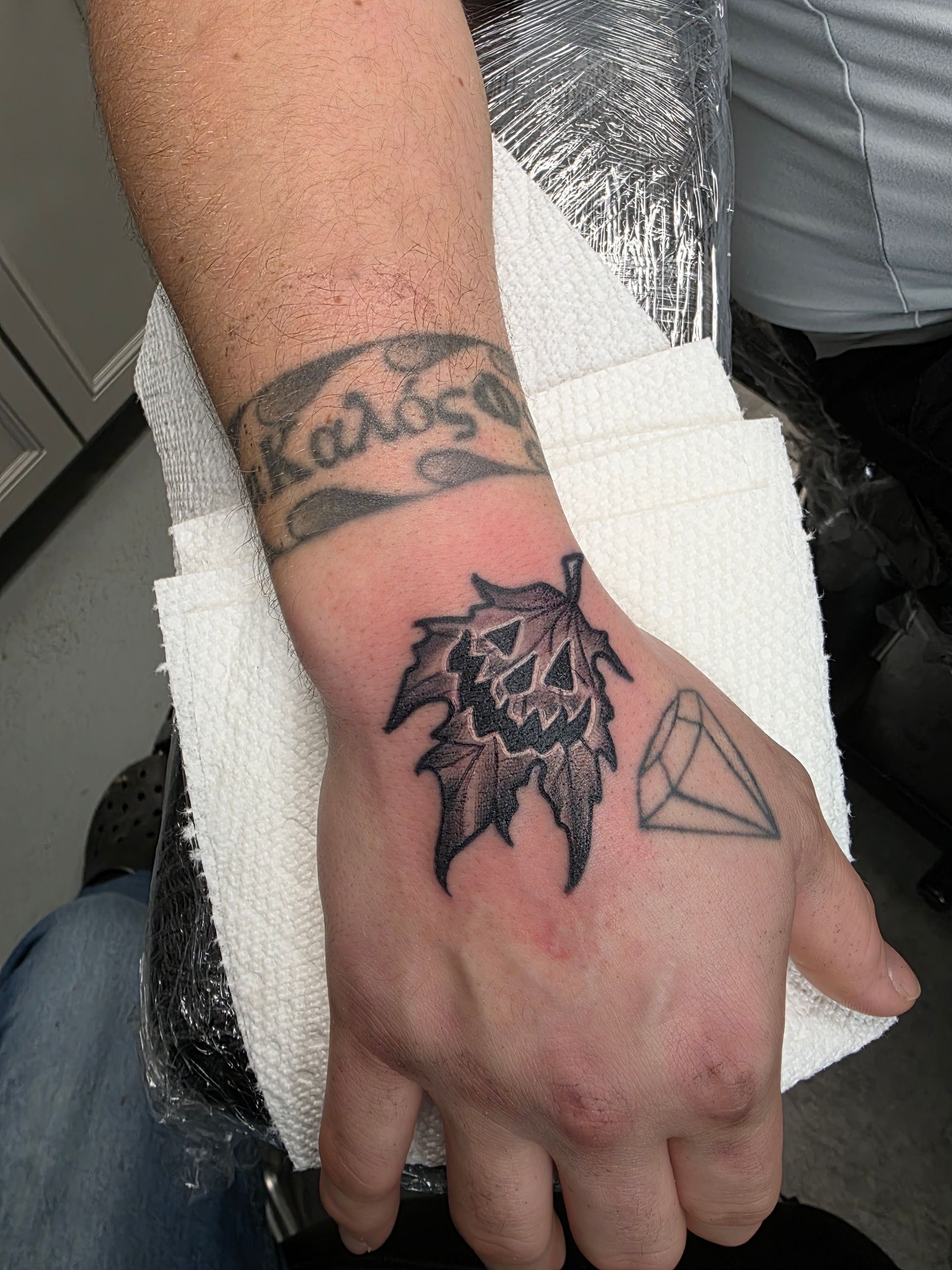 Tattoo of a black leaf on a hand, above a wrist tattoo with Greek text. A geometric symbol is on the hand.