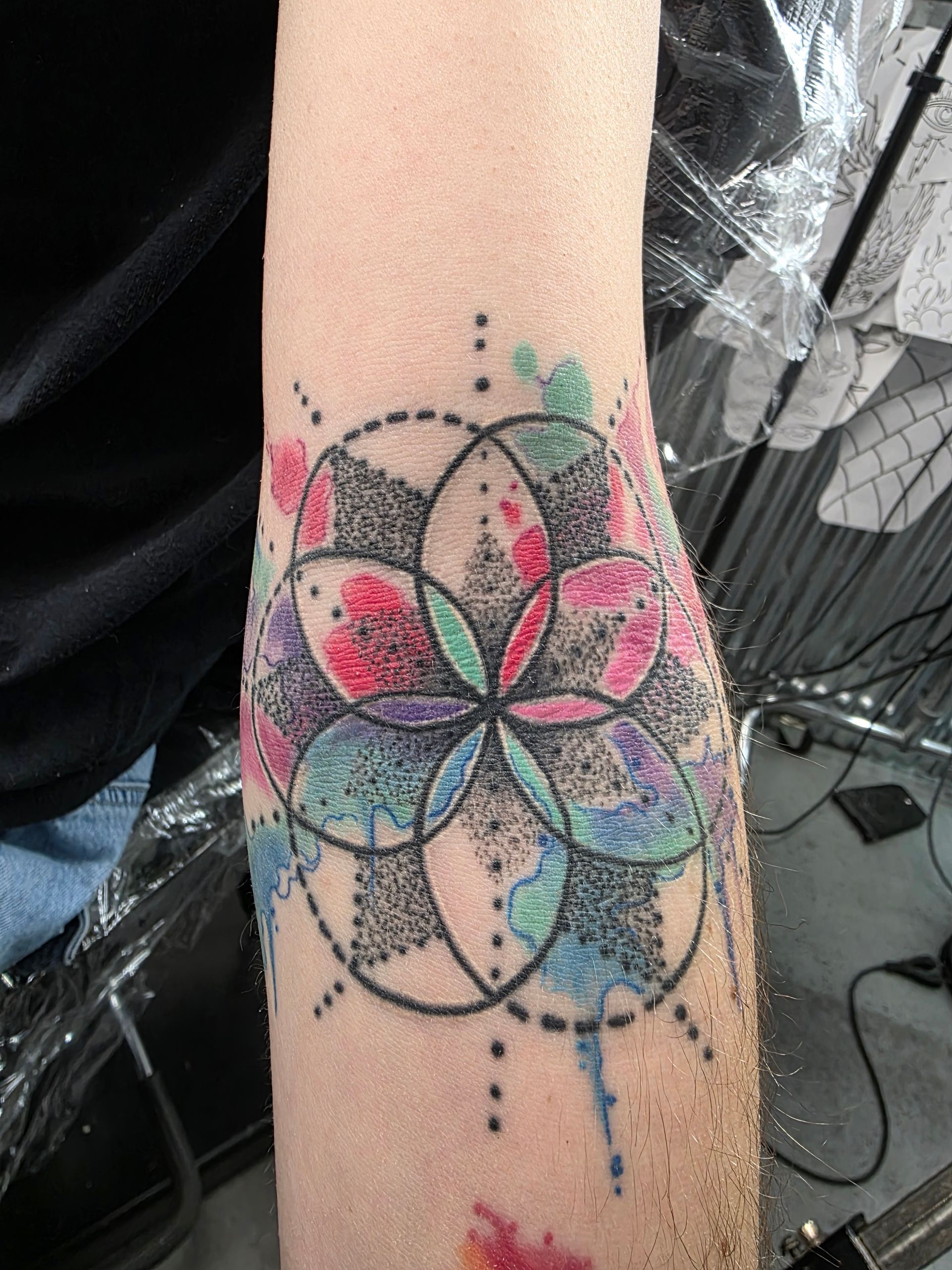 Tattoo of a black dotted outline flower with colorful watercolor effect on arm.