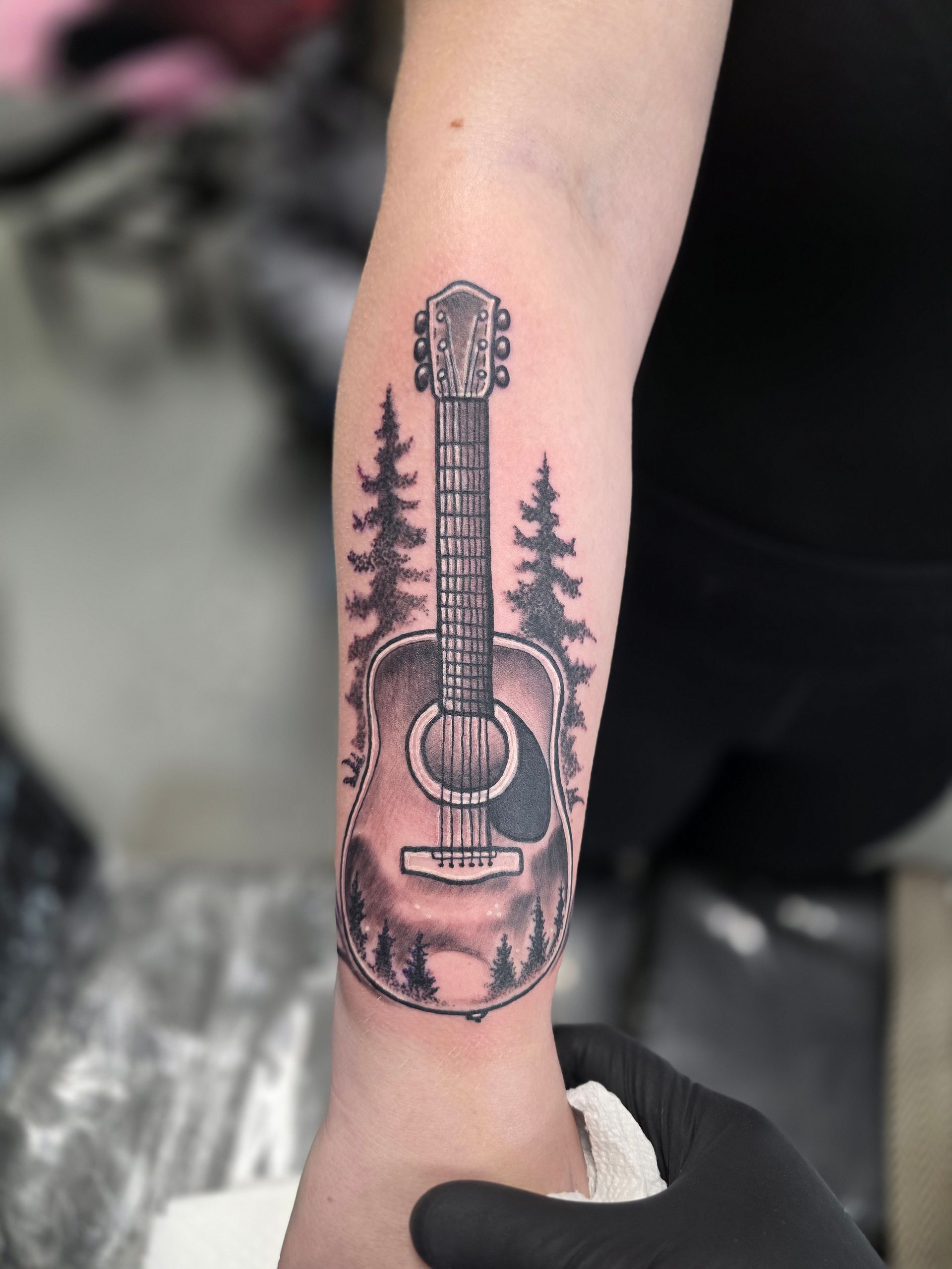 Tattoo of an acoustic guitar and forest scene on a person's forearm, with a hand wearing a black glove.