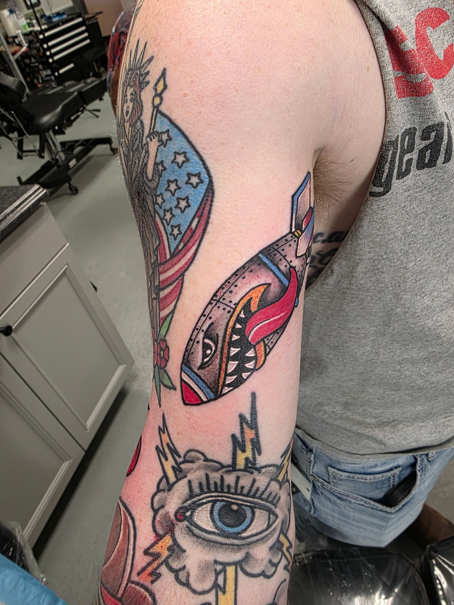 Arm with colorful tattoos including a bomb, flag, eye, and lightning bolts in a tattoo parlor.