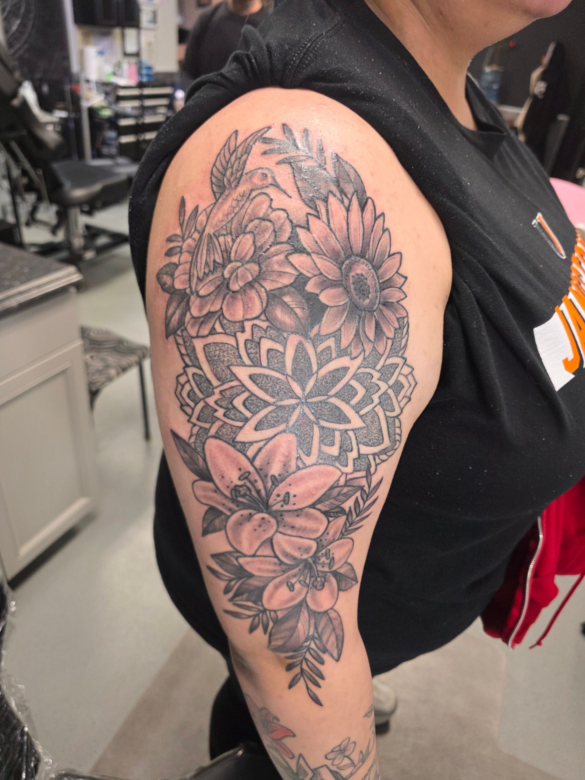 Woman's arm with a detailed black and gray floral tattoo. Includes lilies, sunflowers, and a mandala.