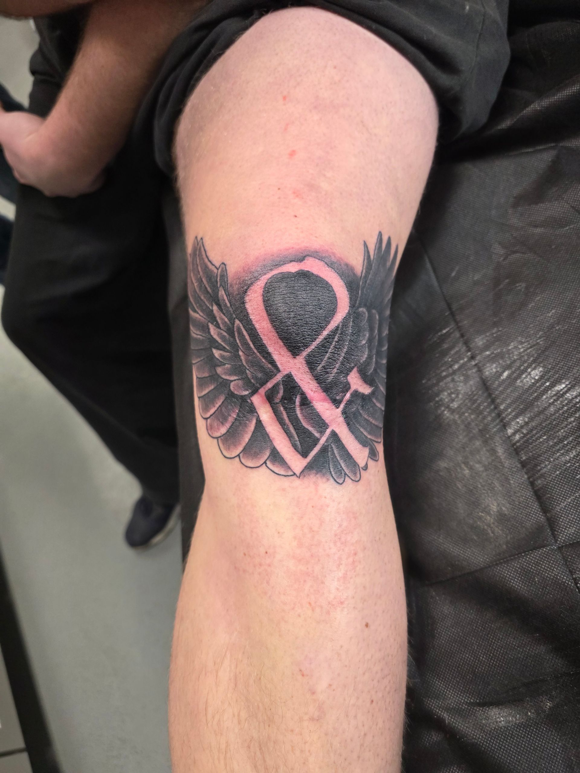 Knee tattoo: winged symbol with two letters, black and pink, set on skin.