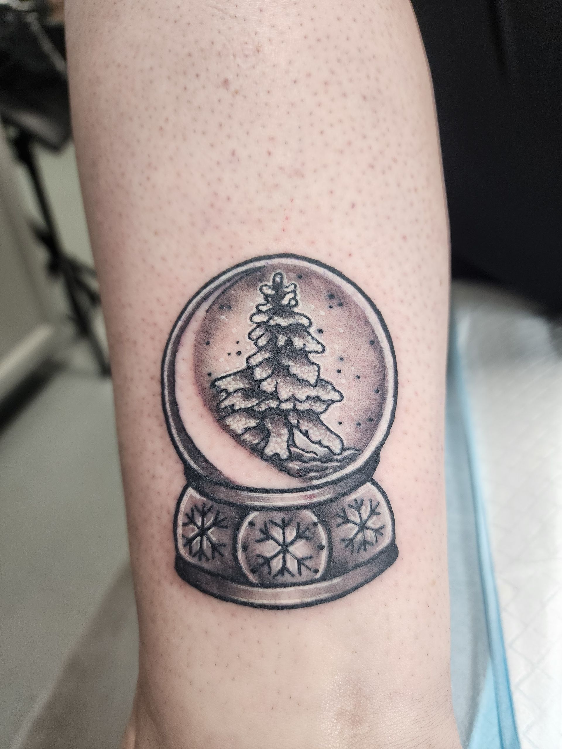 Tattoo of a snow globe with a Christmas tree inside, on an arm. Black and gray shading.