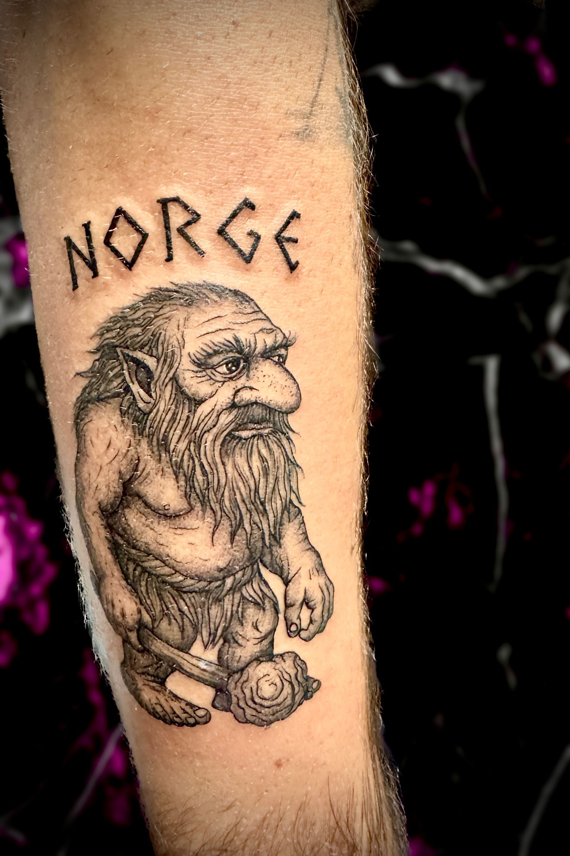 Tattoo of a troll with NORGE above it, on an arm. The troll has a long beard, and it holds a small, round object.