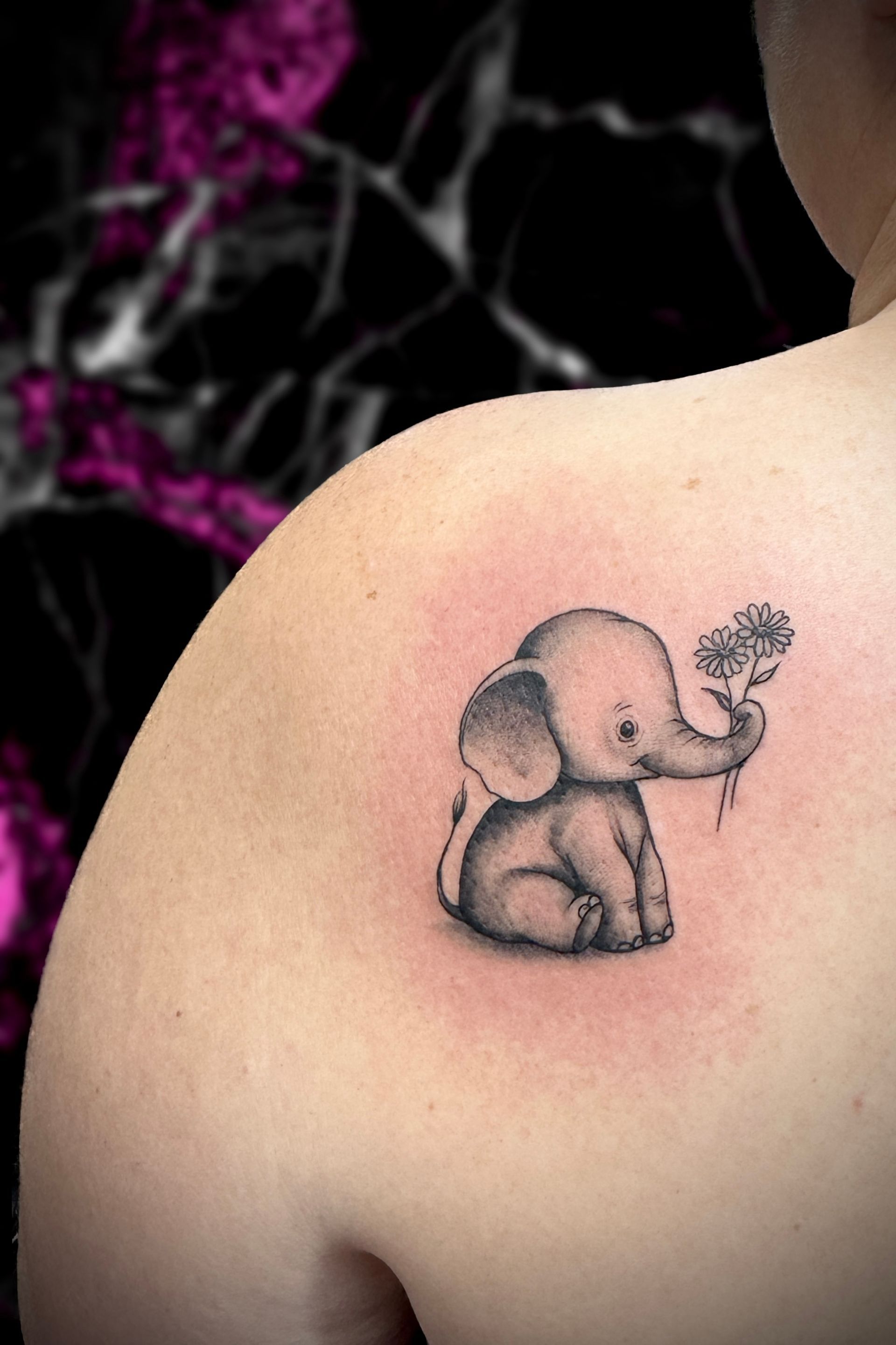 Tattoo of a seated baby elephant holding a small tree on a person's shoulder. Black and gray shading.