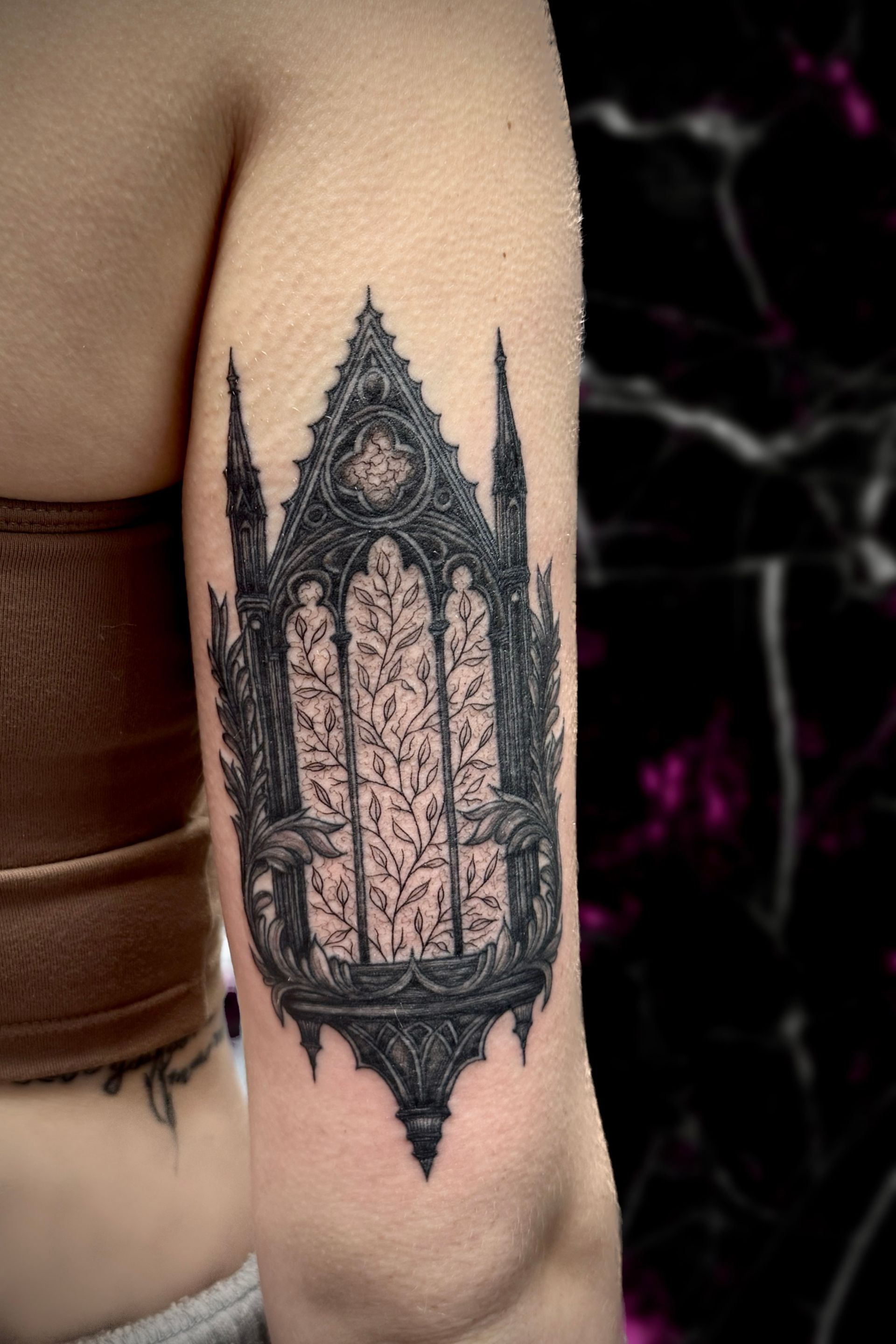 Gothic cathedral window tattoo on a person's upper arm, intricate black design with foliage inside.