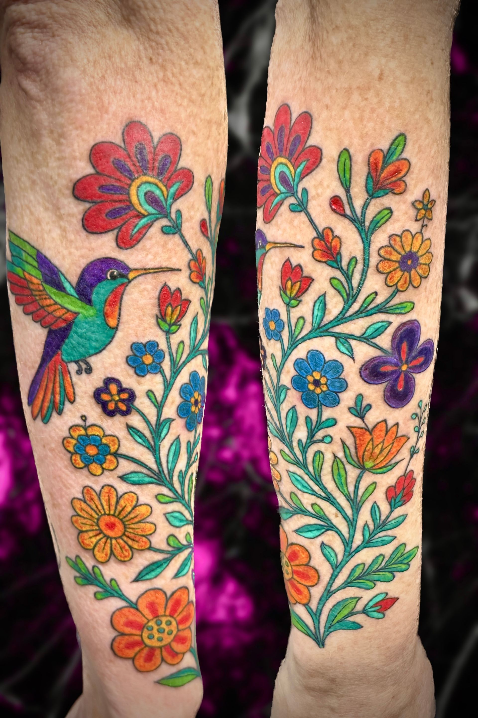 Colorful floral tattoo on forearms, includes a hummingbird and various flowers.