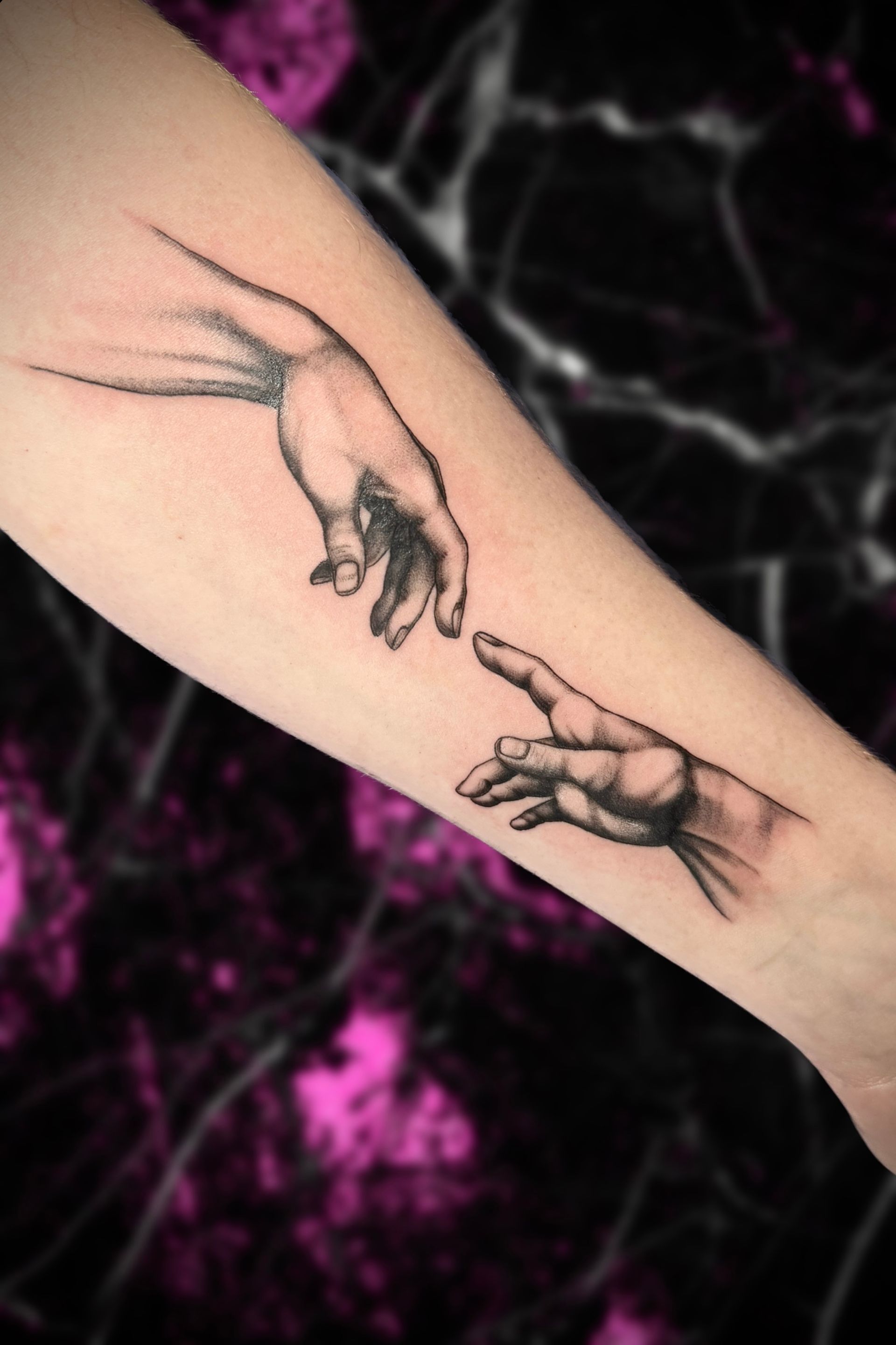Tattoo of two hands, mimicking Michelangelo's Creation of Adam, on an arm, black and white.