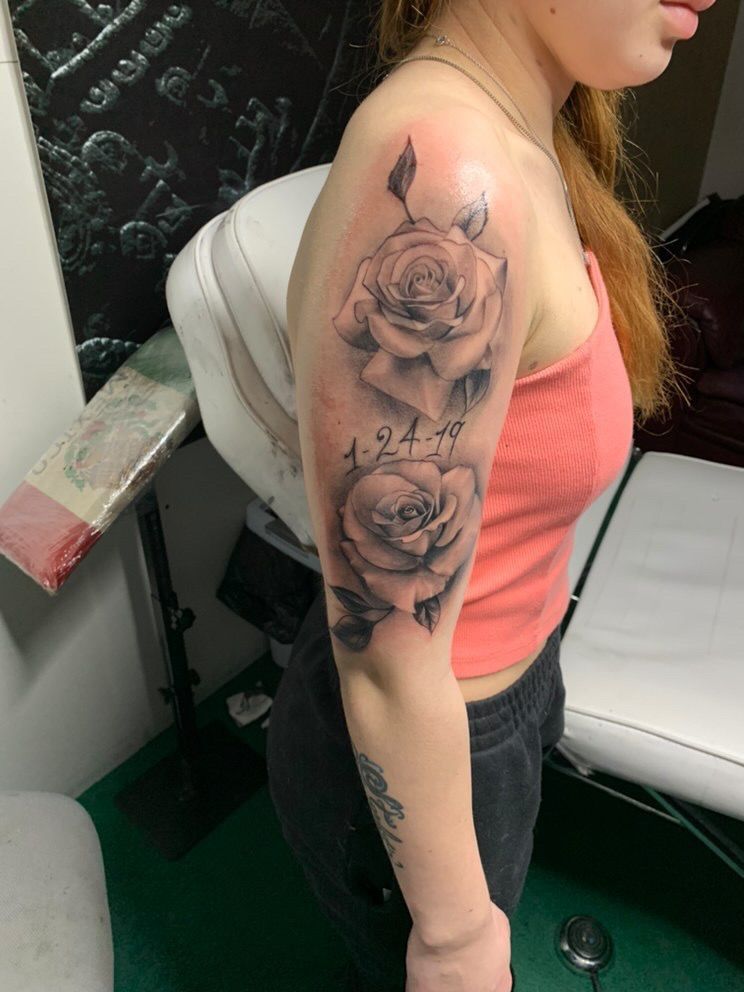 Woman with rose tattoos on her arm, wearing a pink tank top and black pants, in a tattoo parlor.