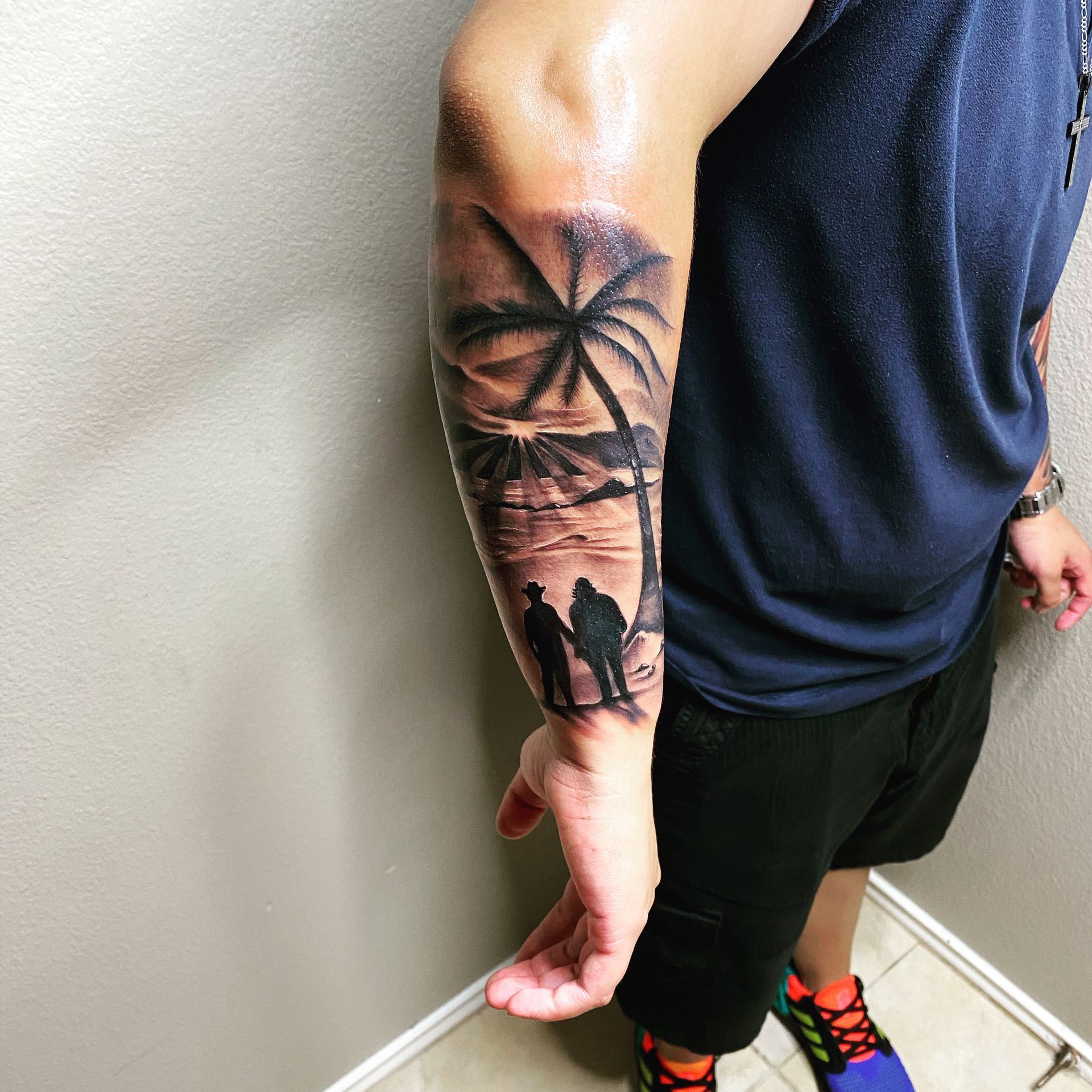 Black ink tattoo on forearm of a beach scene with palm tree, couple holding hands, and sunset.