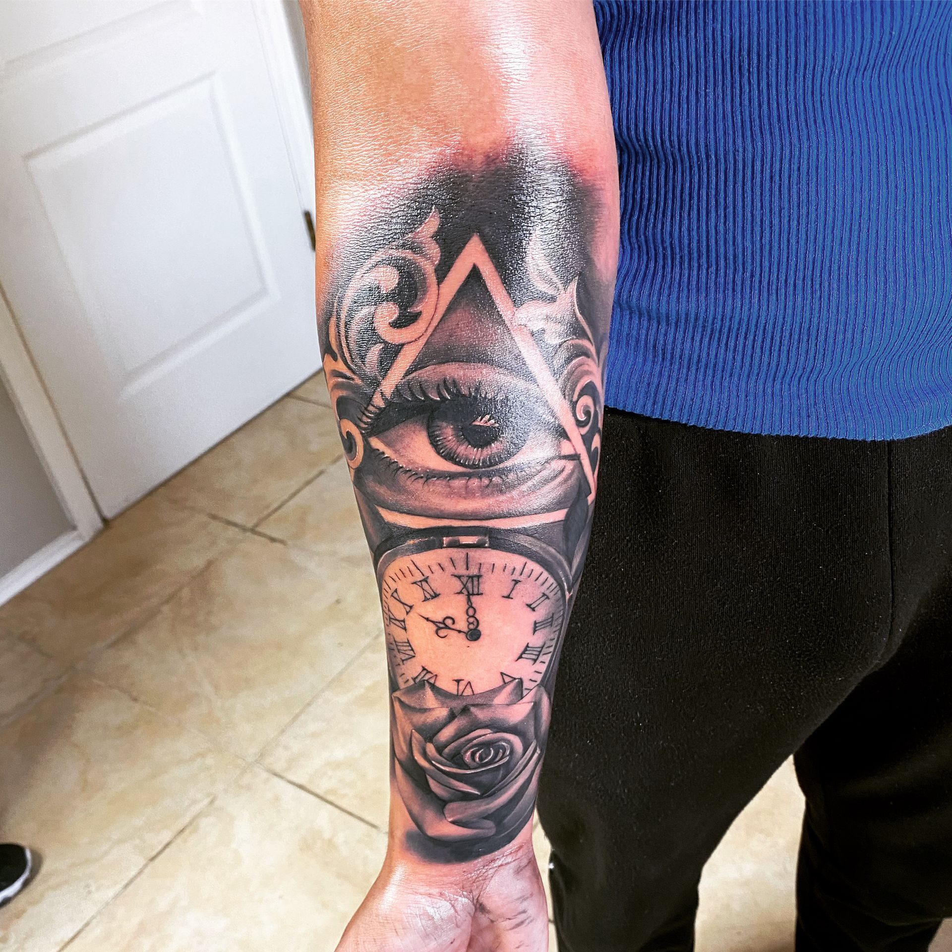 Tattoo of an eye within a triangle, clock, and rose on a person's arm. Black and gray.