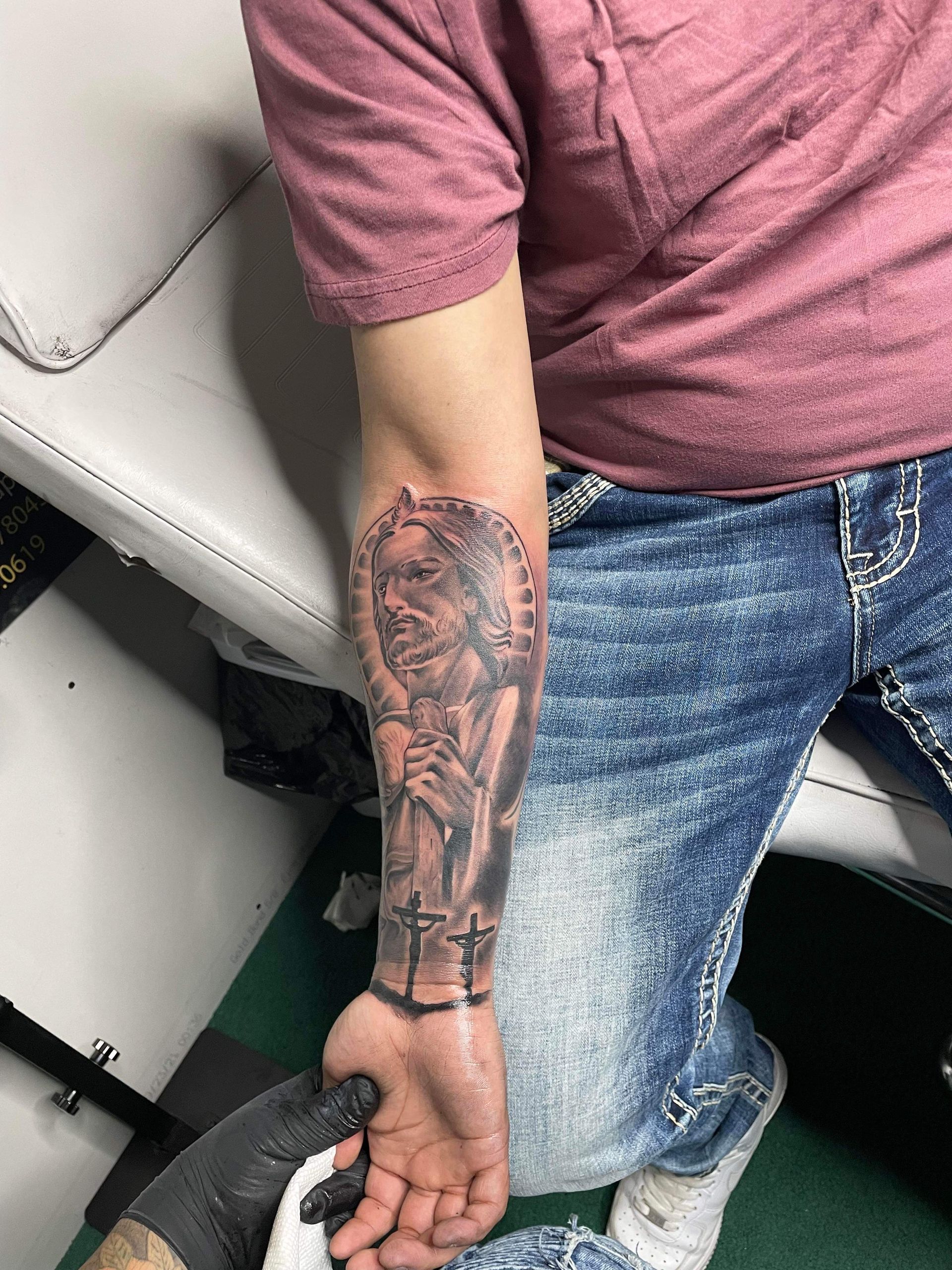 Person's arm with a tattoo of Jesus wearing a crown, with crosses below. The person is wearing jeans and a pink shirt.