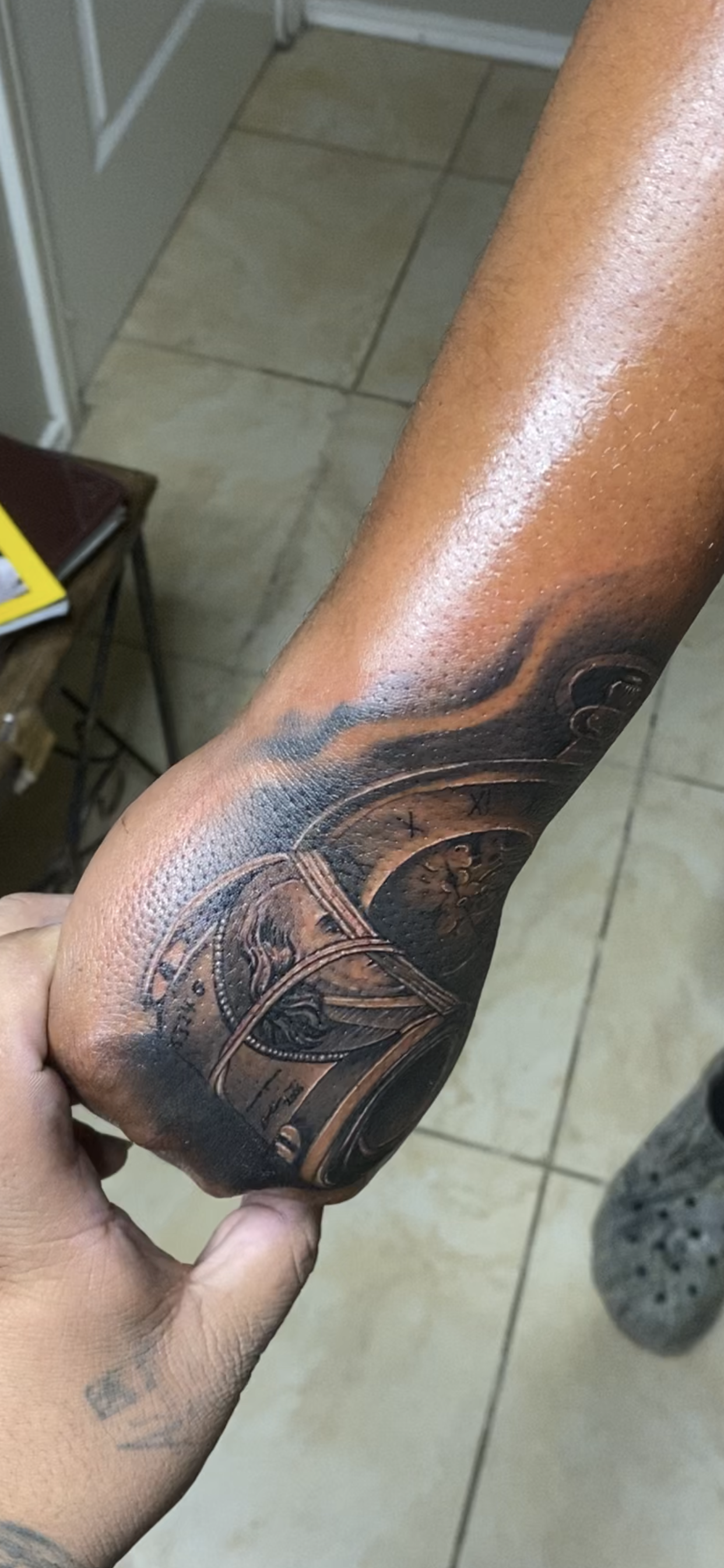 Arm with tattoo, possibly new, near elbow. Holding hand present. Indoors, tiled floor.