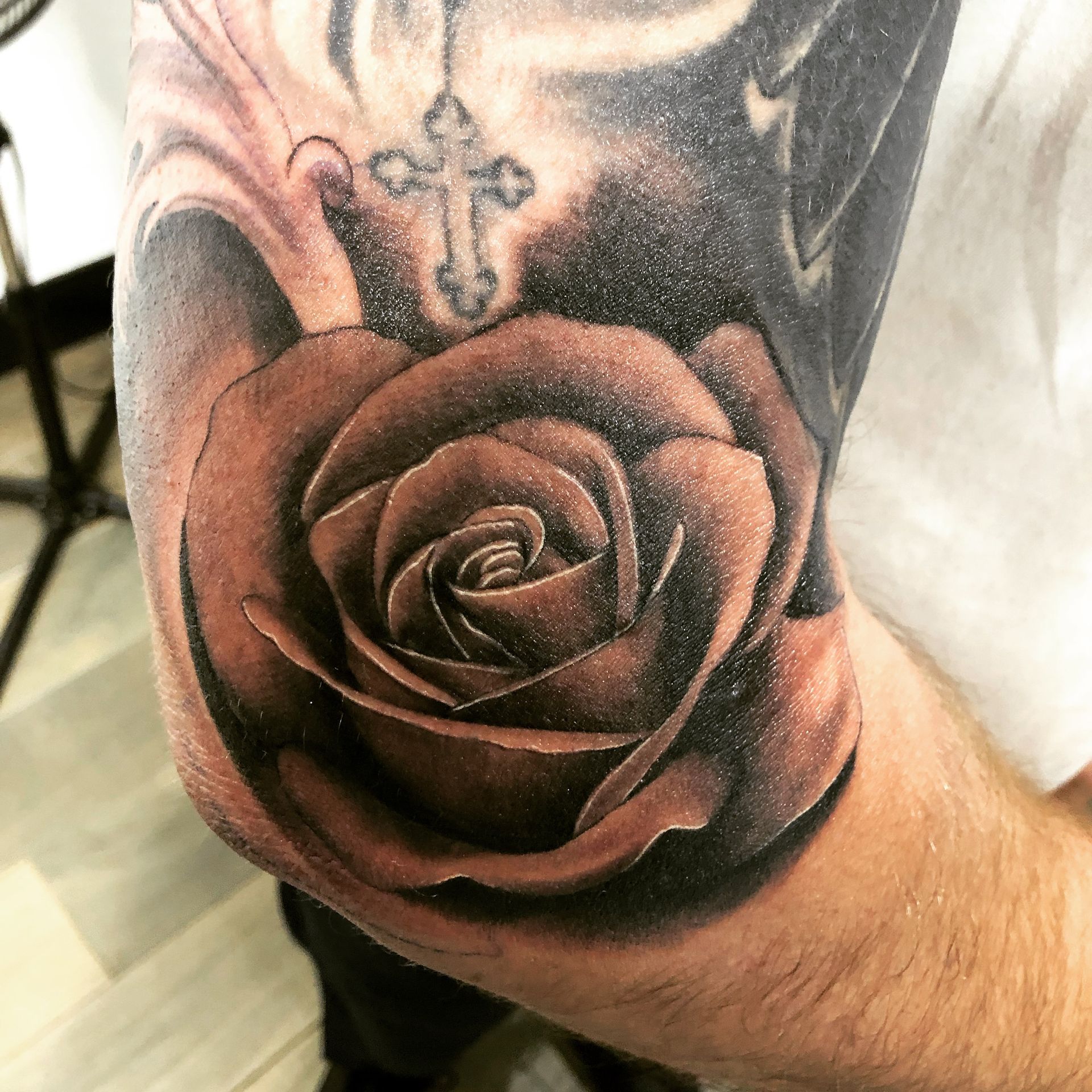 Rose tattoo in black and brown on a person's arm, with a cross above the rose.