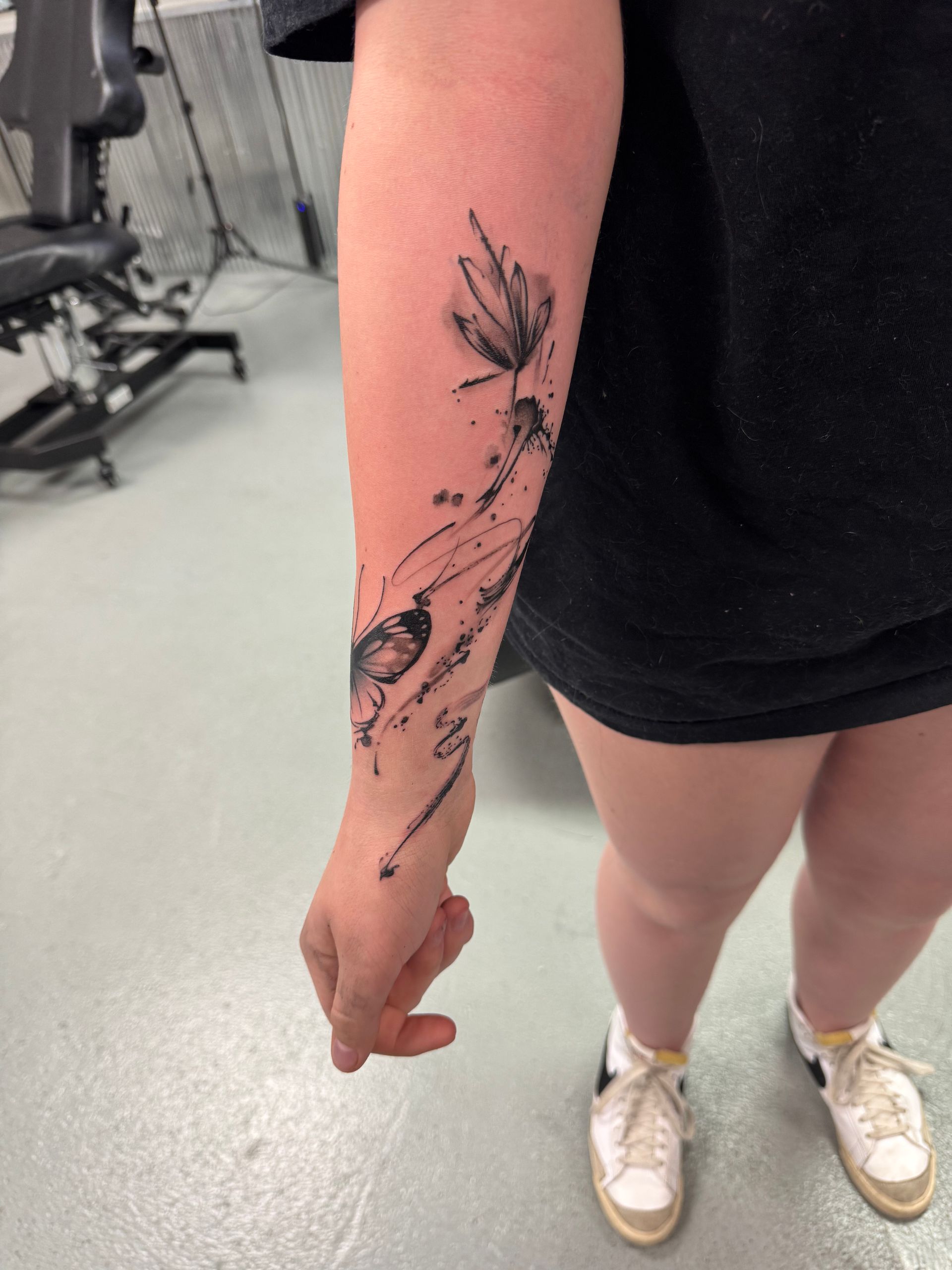Tattoo of abstract floral design on a person's forearm; person standing, wearing black shirt and white sneakers.