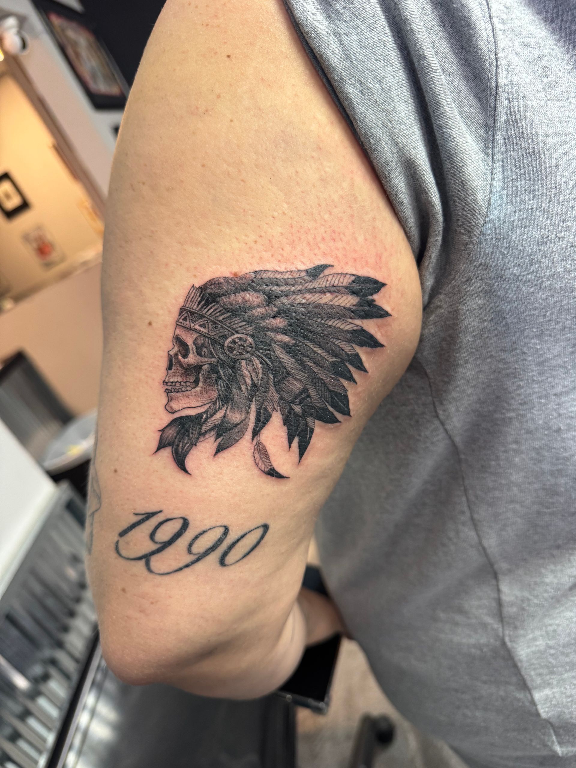 Tattoo of skull wearing a Native American headdress, with the year 1990 below, on a person's arm.