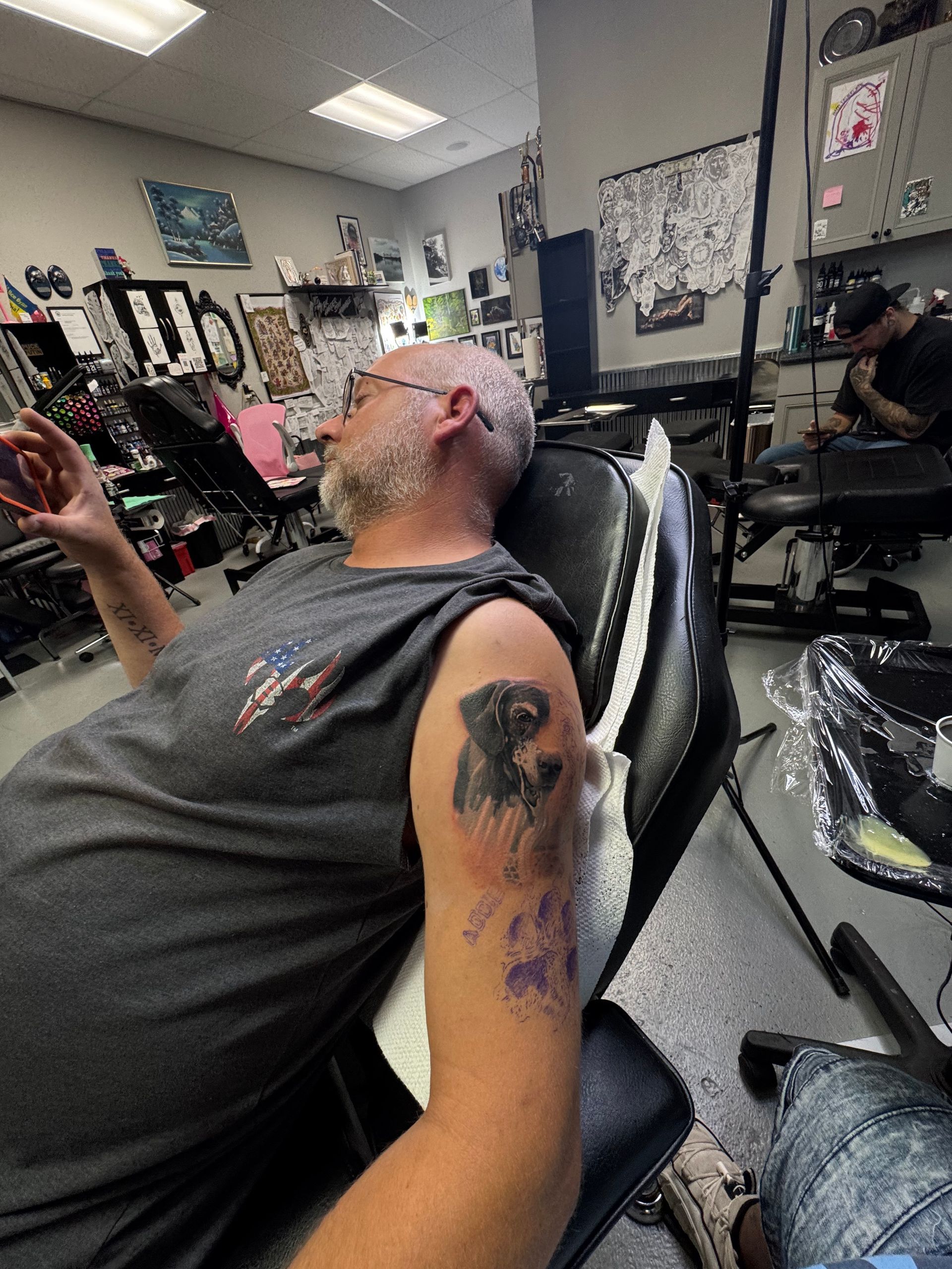 Man getting a dog tattoo on his arm at a tattoo parlor.  He's wearing a gray shirt and sitting in a black chair.