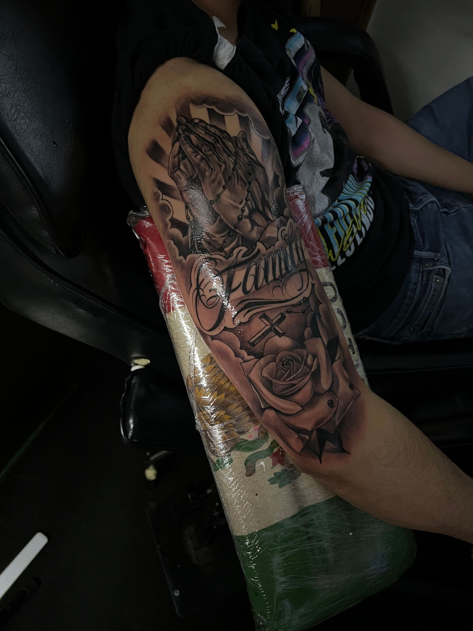 Arm tattoo: Praying hands, roses, and text, wrapped in plastic. Person seated.