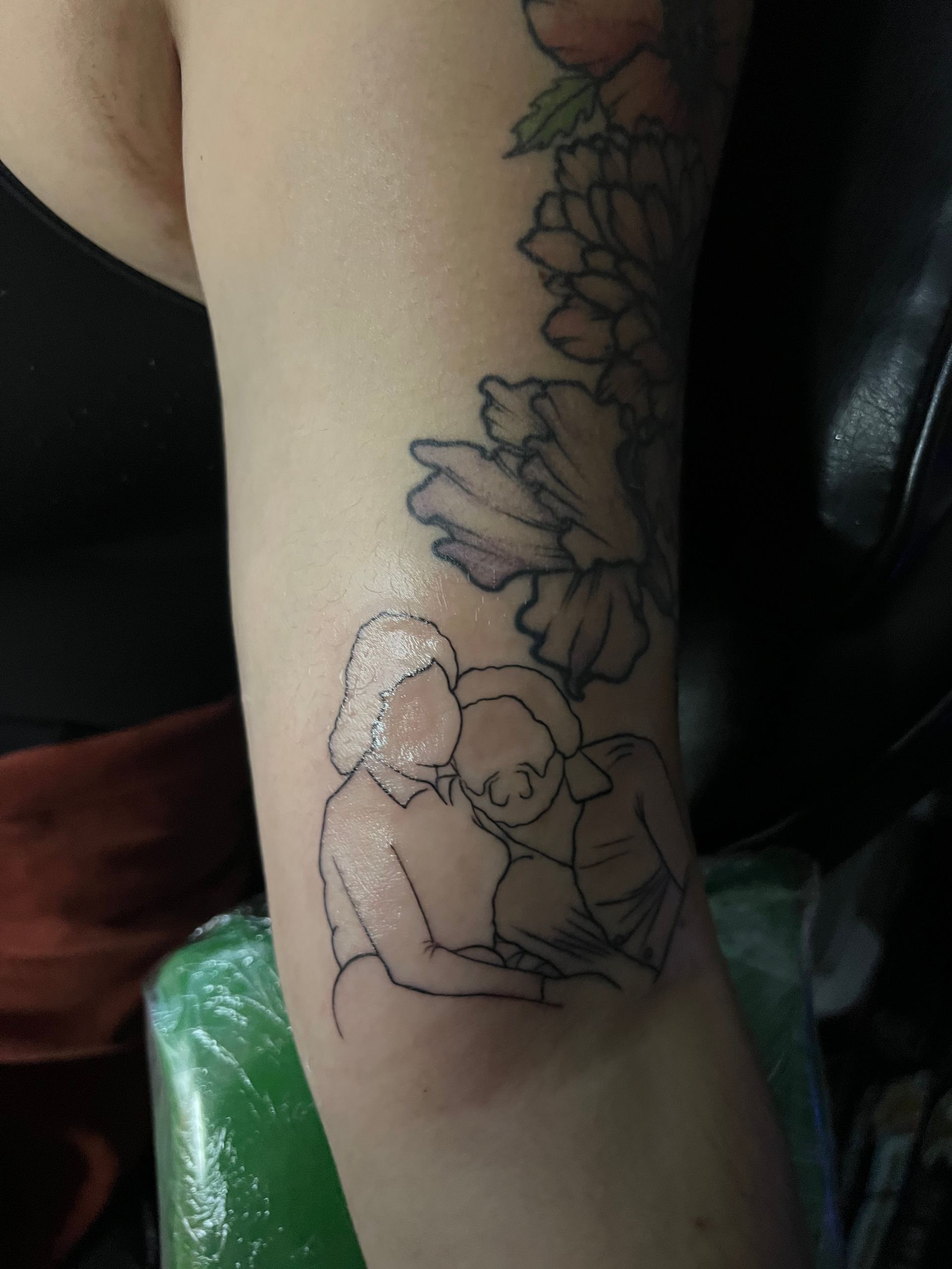 Tattoo of two figures, a person hugging another while reading, with flower outline on arm.