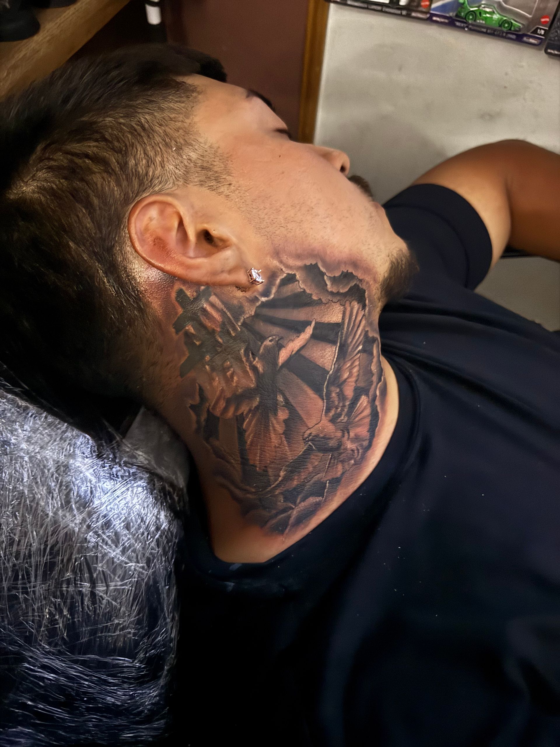 Man with a neck tattoo of a rising sun design, dark hair, wearing a black shirt, lying down.