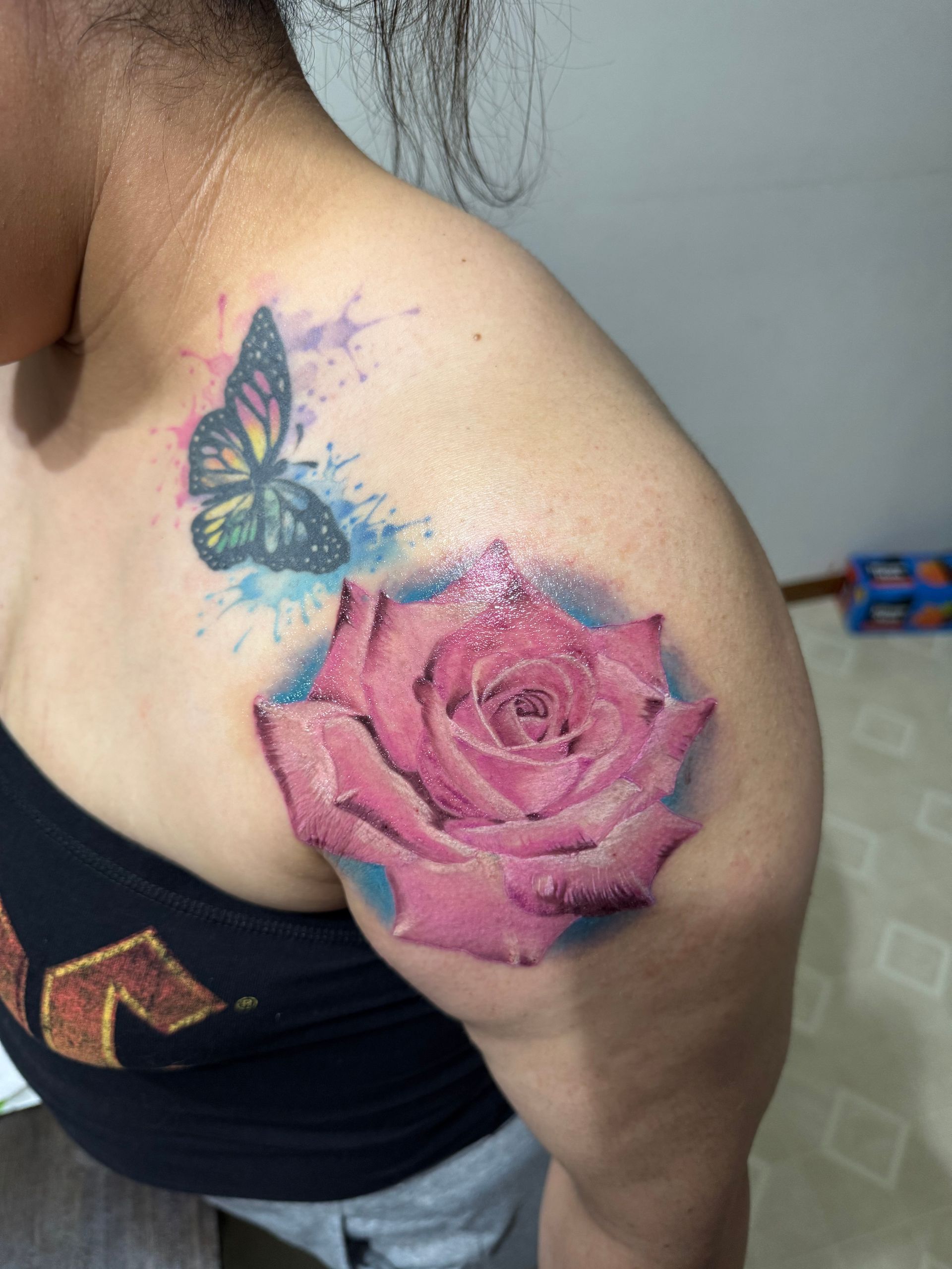 Woman's shoulder tattoo featuring a pink rose and a butterfly with blue watercolor splatters.