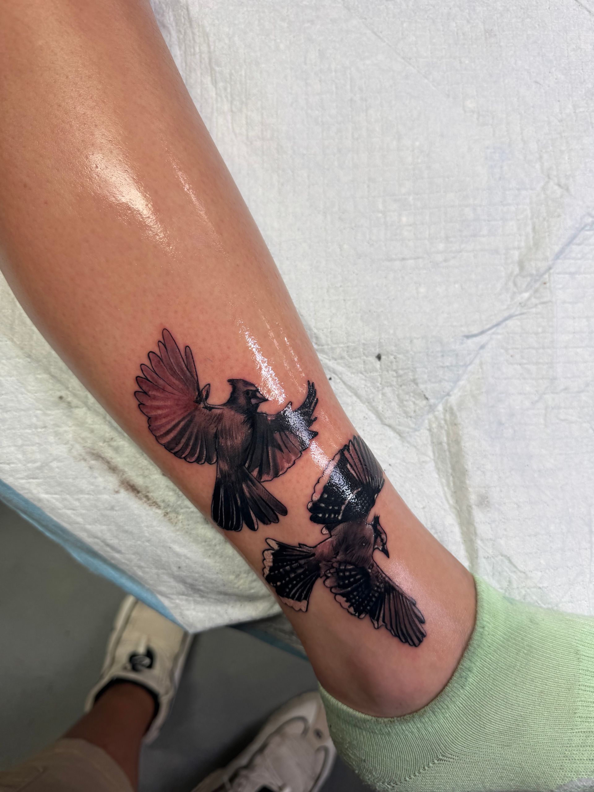 Tattoo of three birds in flight on a leg; one with a red wing, on light skin, next to a green sock and shoe.