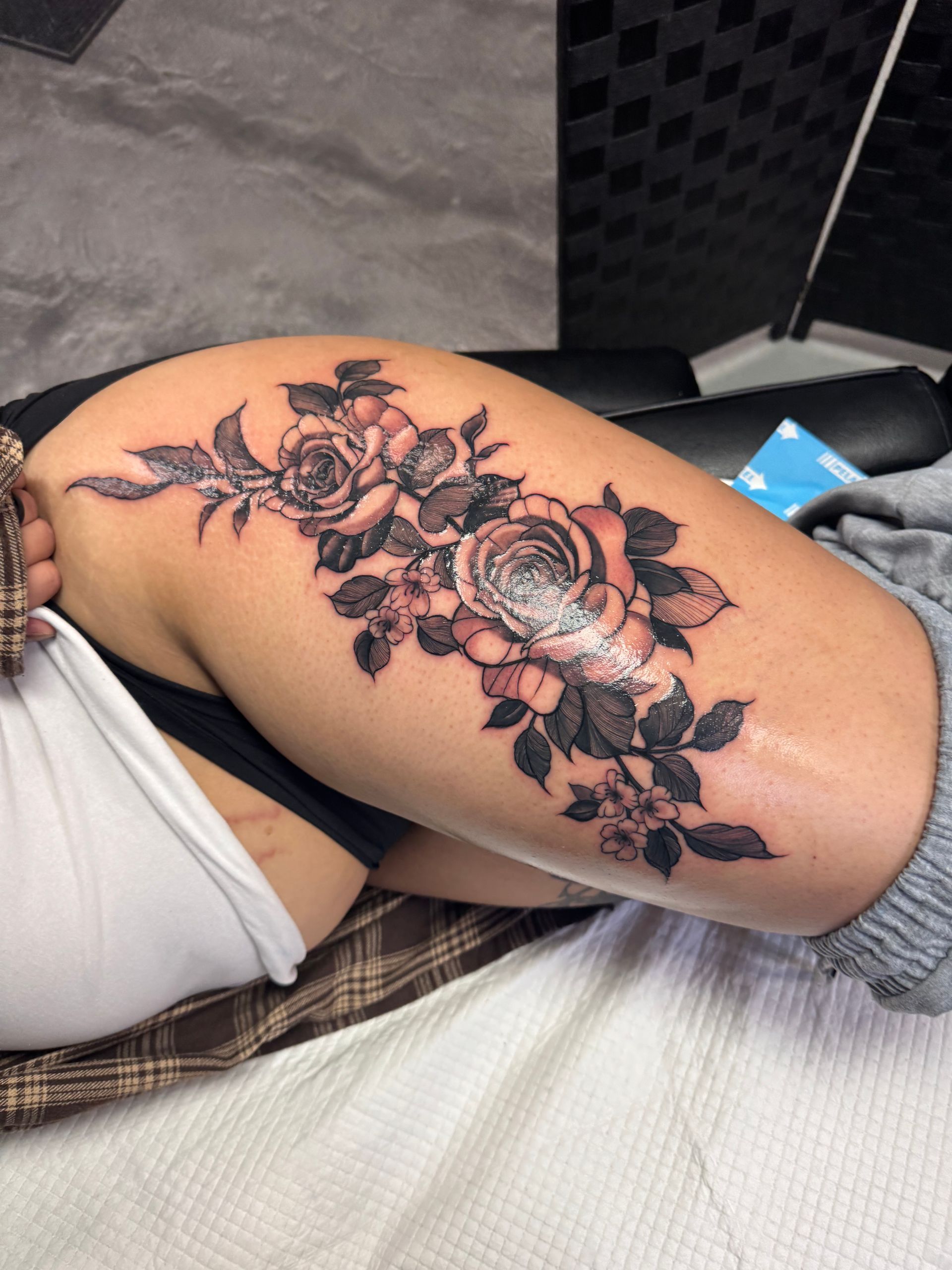 Black and gray floral tattoo on a person's thigh. Roses and leaves.