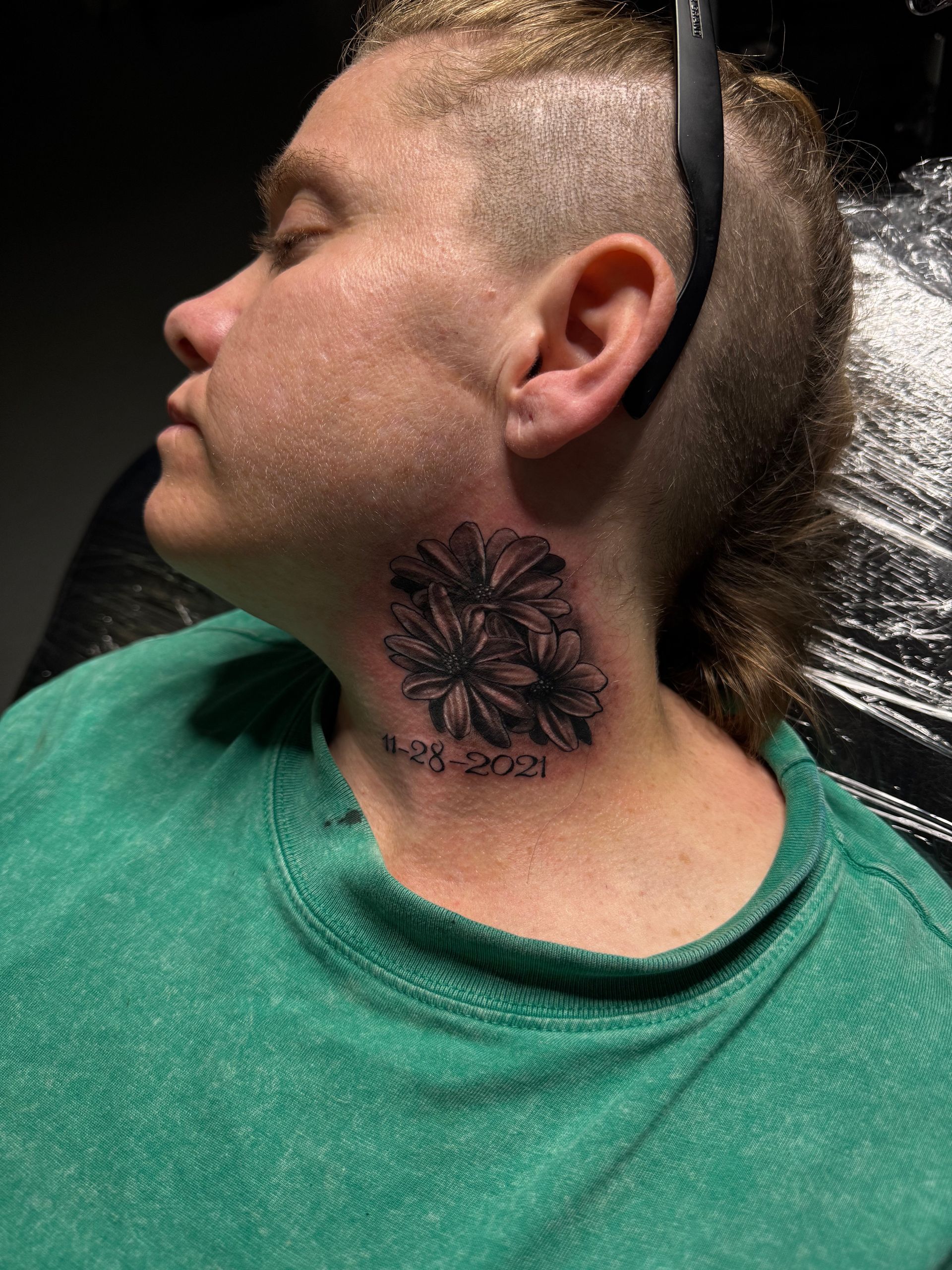 Woman with a floral neck tattoo, shaded, and date 11-28-2022. Wearing green shirt, hair cut short.