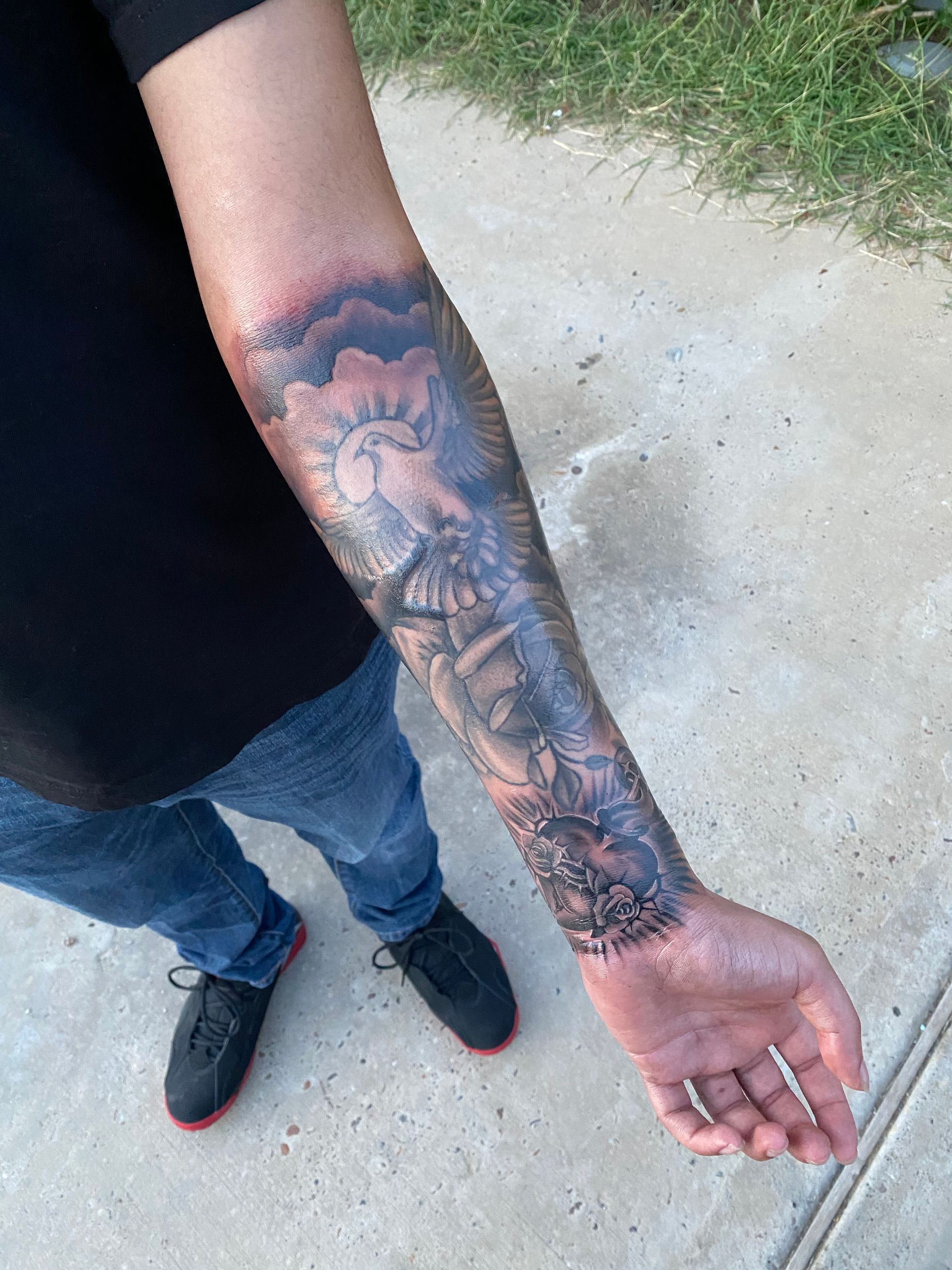 Man's forearm with a detailed black and gray tattoo of a sun, clouds, and figures. He wears jeans and black shoes.