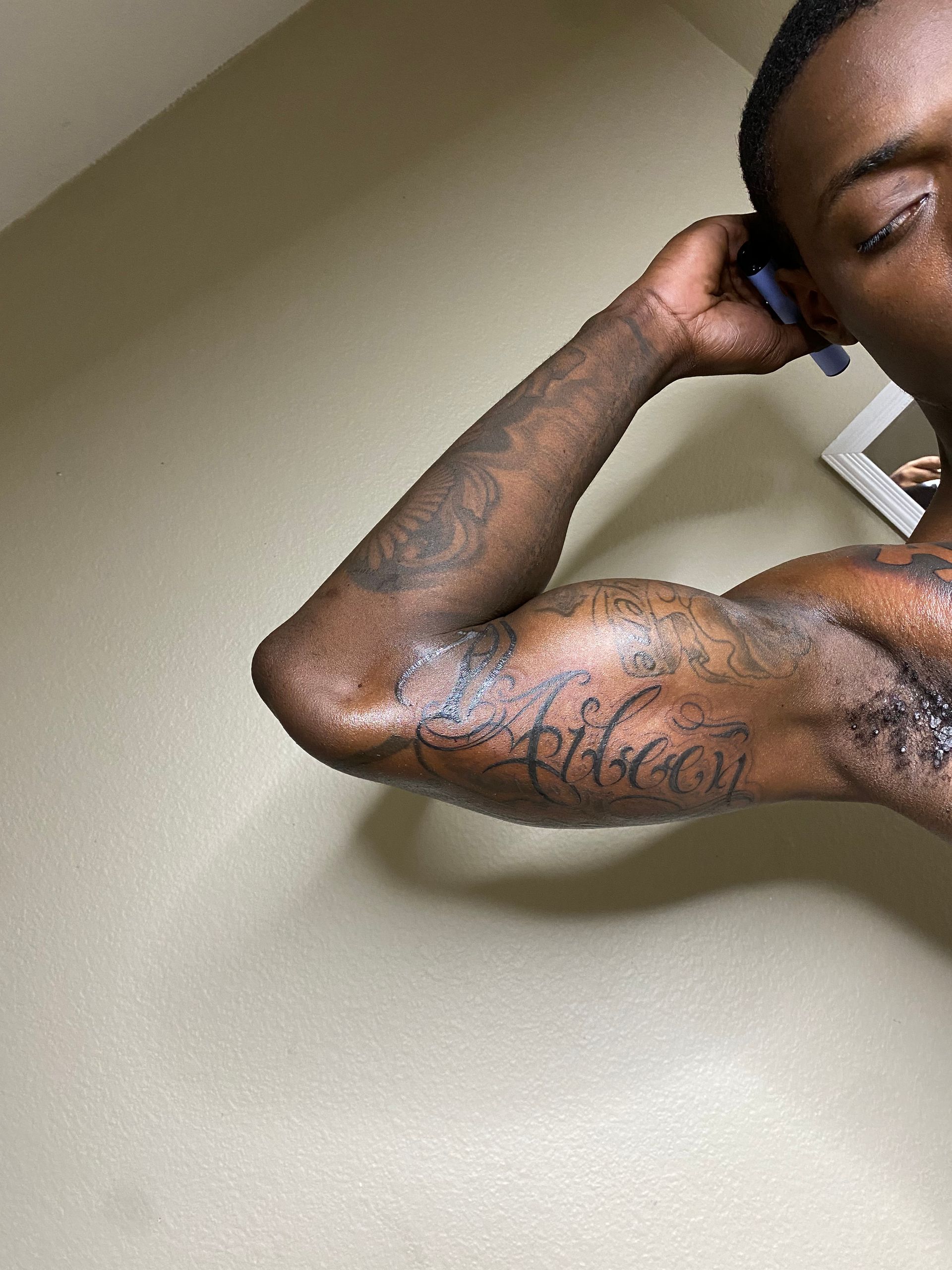 Man flexing arm, showing tattoos and muscles, dark skin, holding phone, indoors.