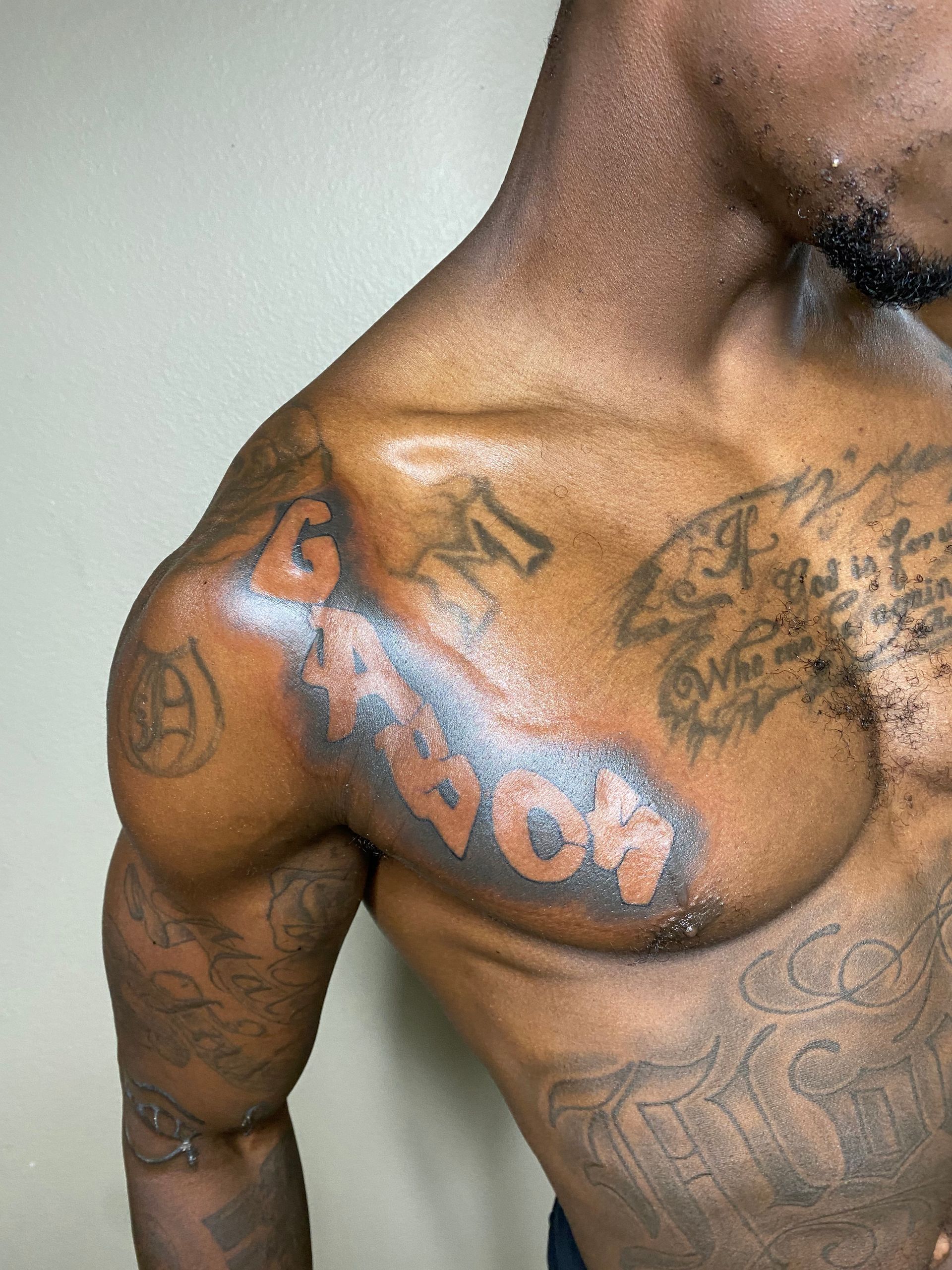 Man with tattoos on chest and arm, flexing shoulder. Dark skin, glistening.