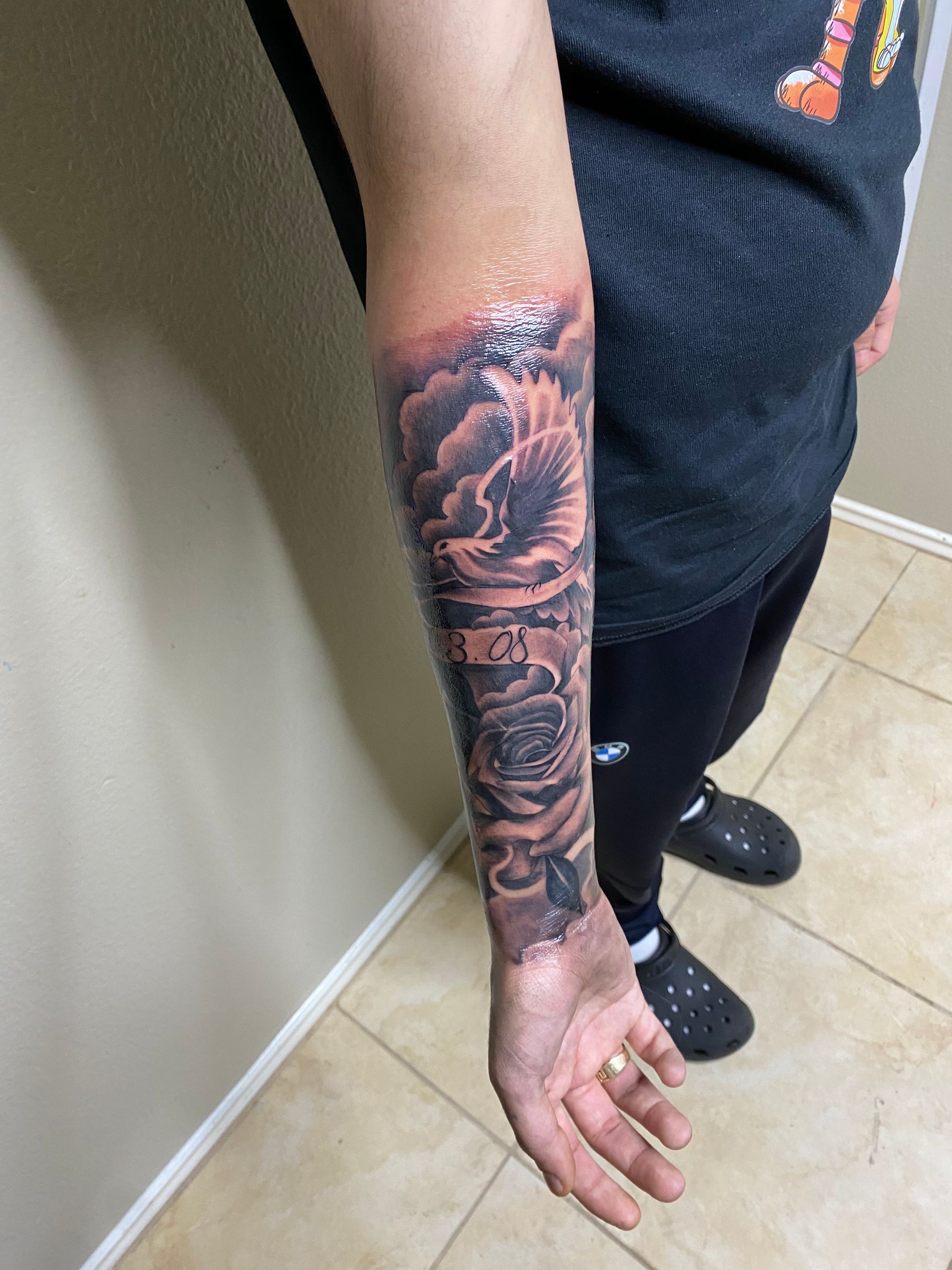 Tattoo of a dove and rose on a person's forearm, in black ink with clouds.