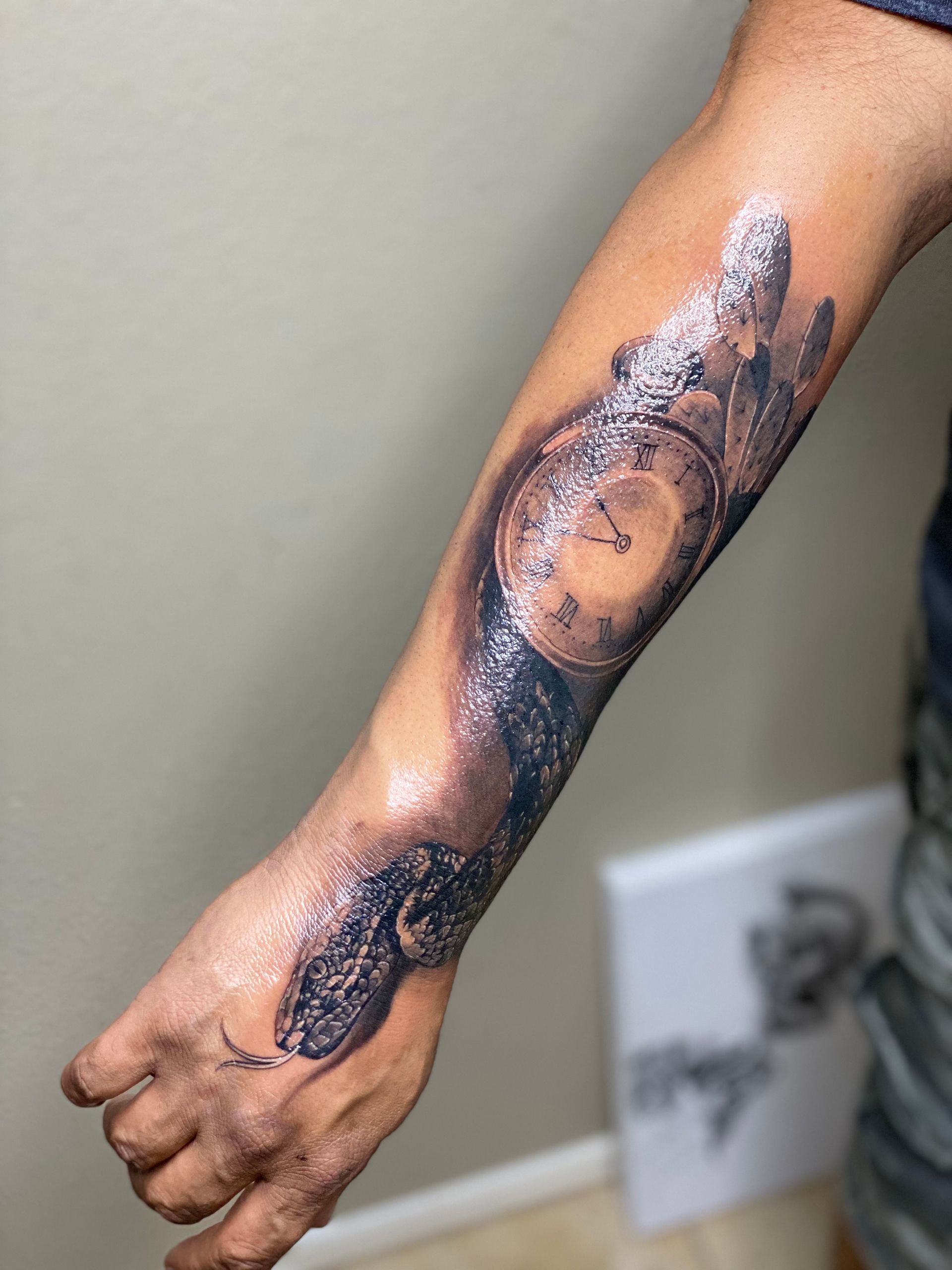 Tattoo of a pocket watch and snake on arm, black and gray ink, against a tan wall.