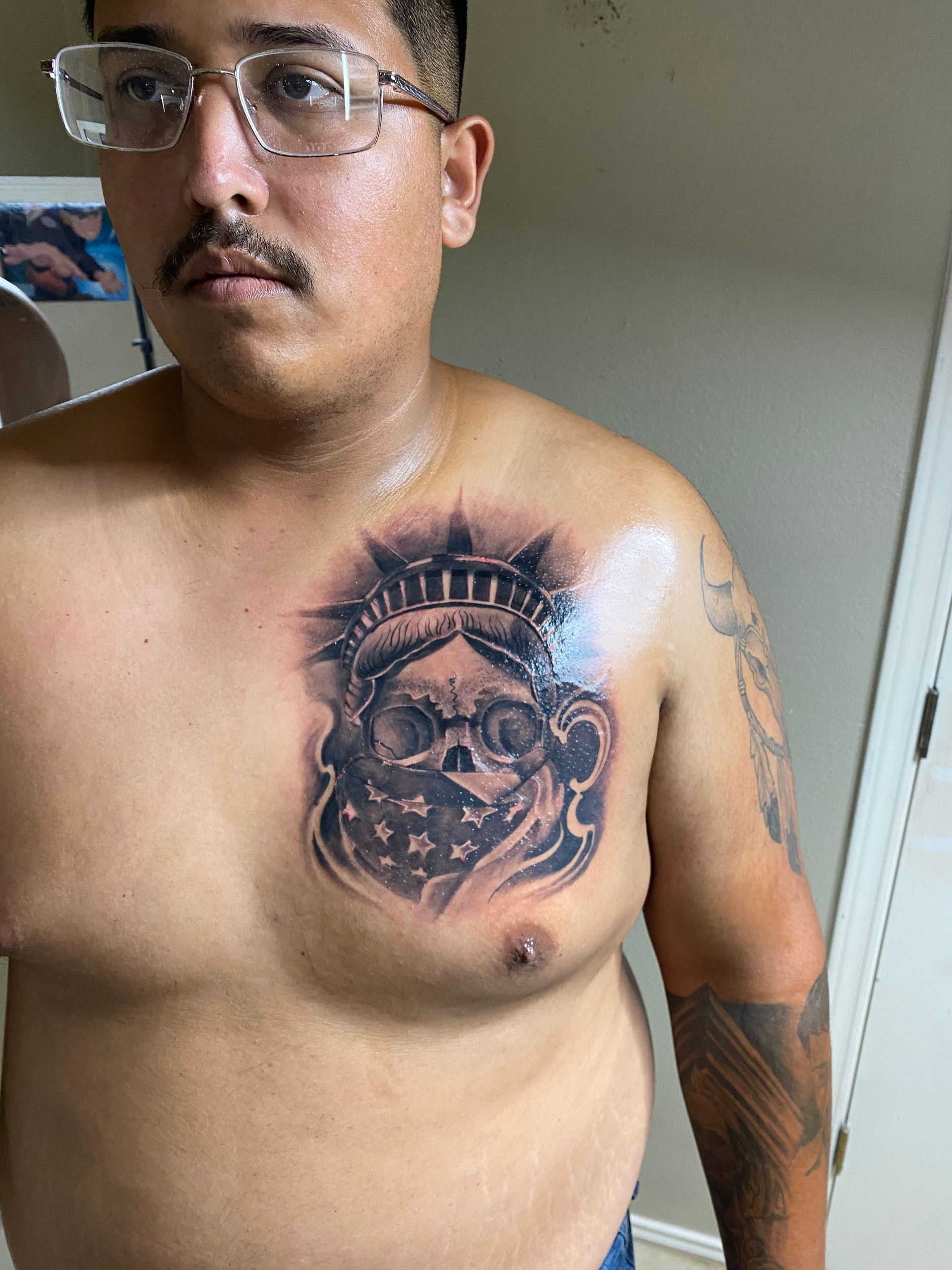 Man with chest tattoo of skull wearing Statue of Liberty crown, and bandana.