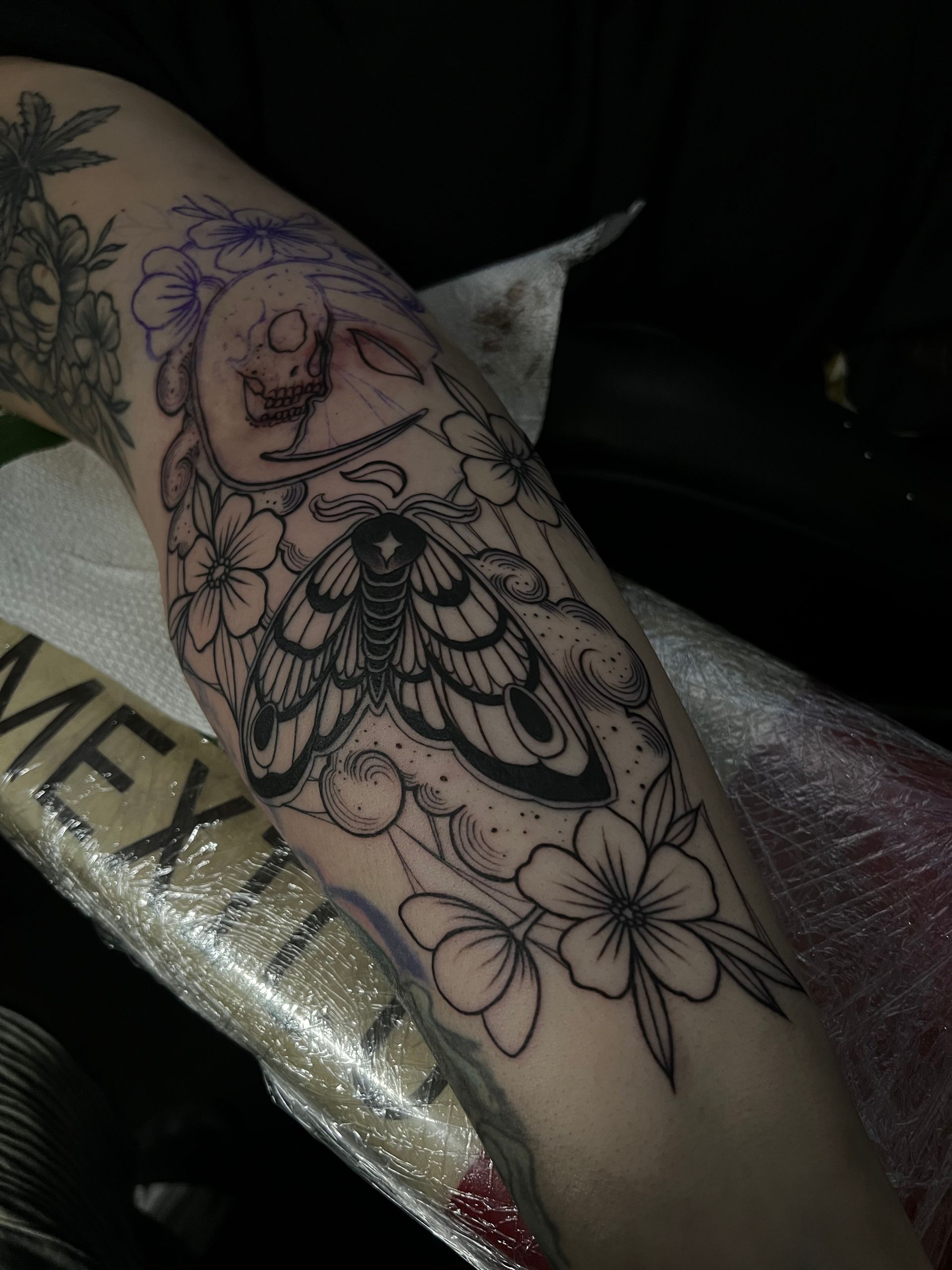Tattoo of a moth with floral details and a skull on an arm. Black ink design, in progress.