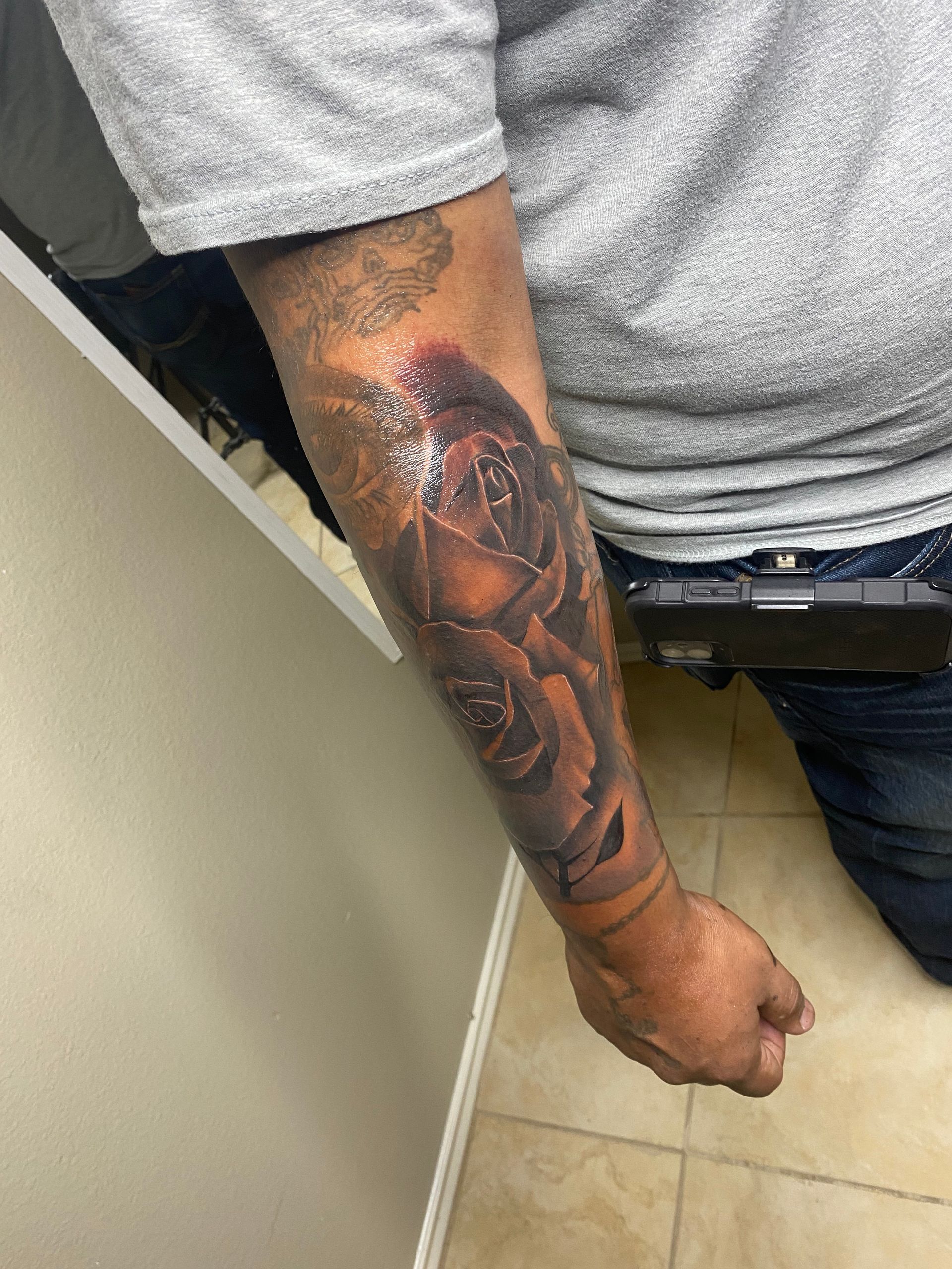 Person's arm with a detailed black and grey rose tattoo. They wear a grey shirt and jeans, standing by a wall.