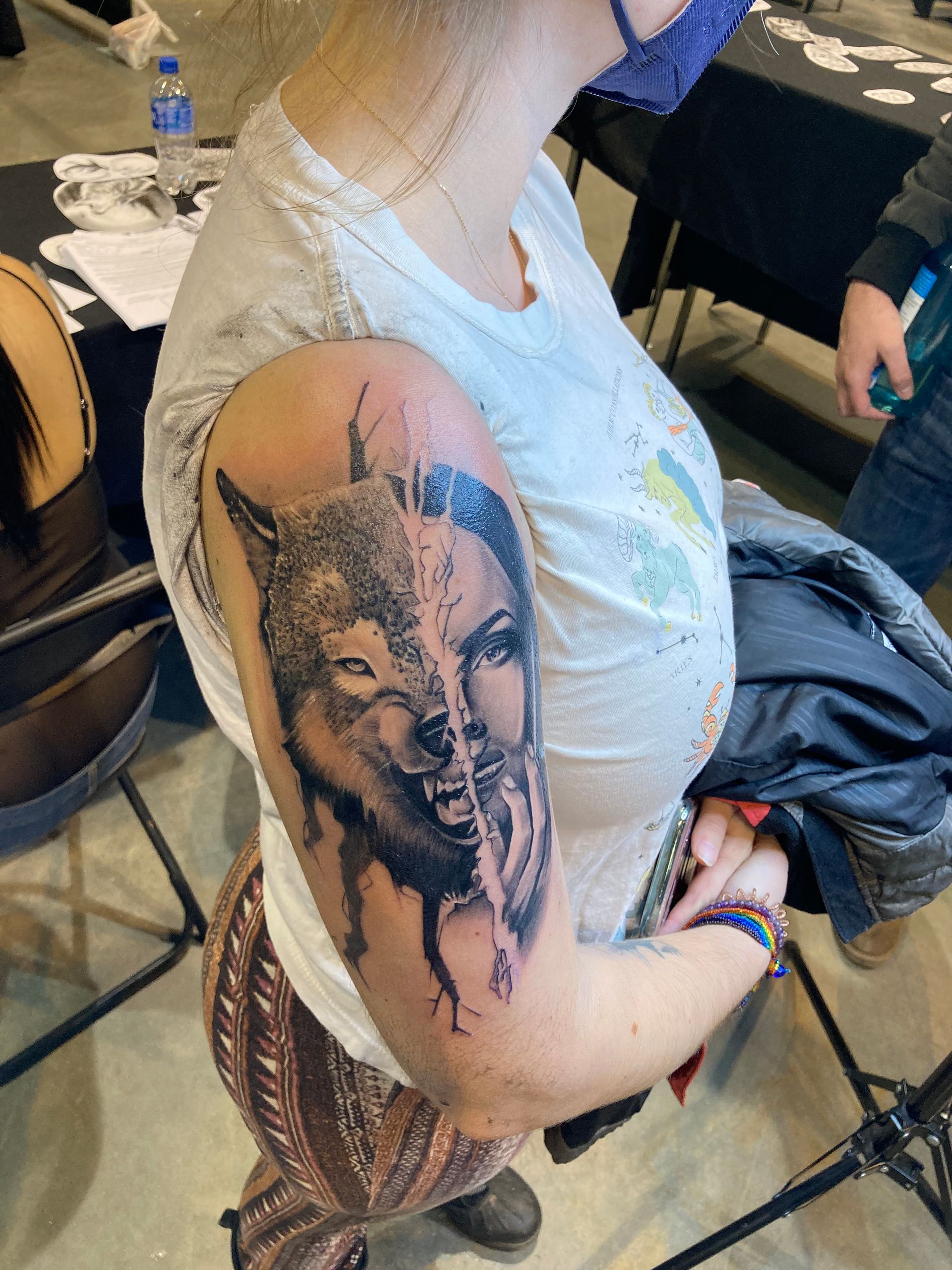 Woman with a wolf and woman face tattoo on her upper arm at a tattoo parlor. She wears a white shirt and patterned pants.