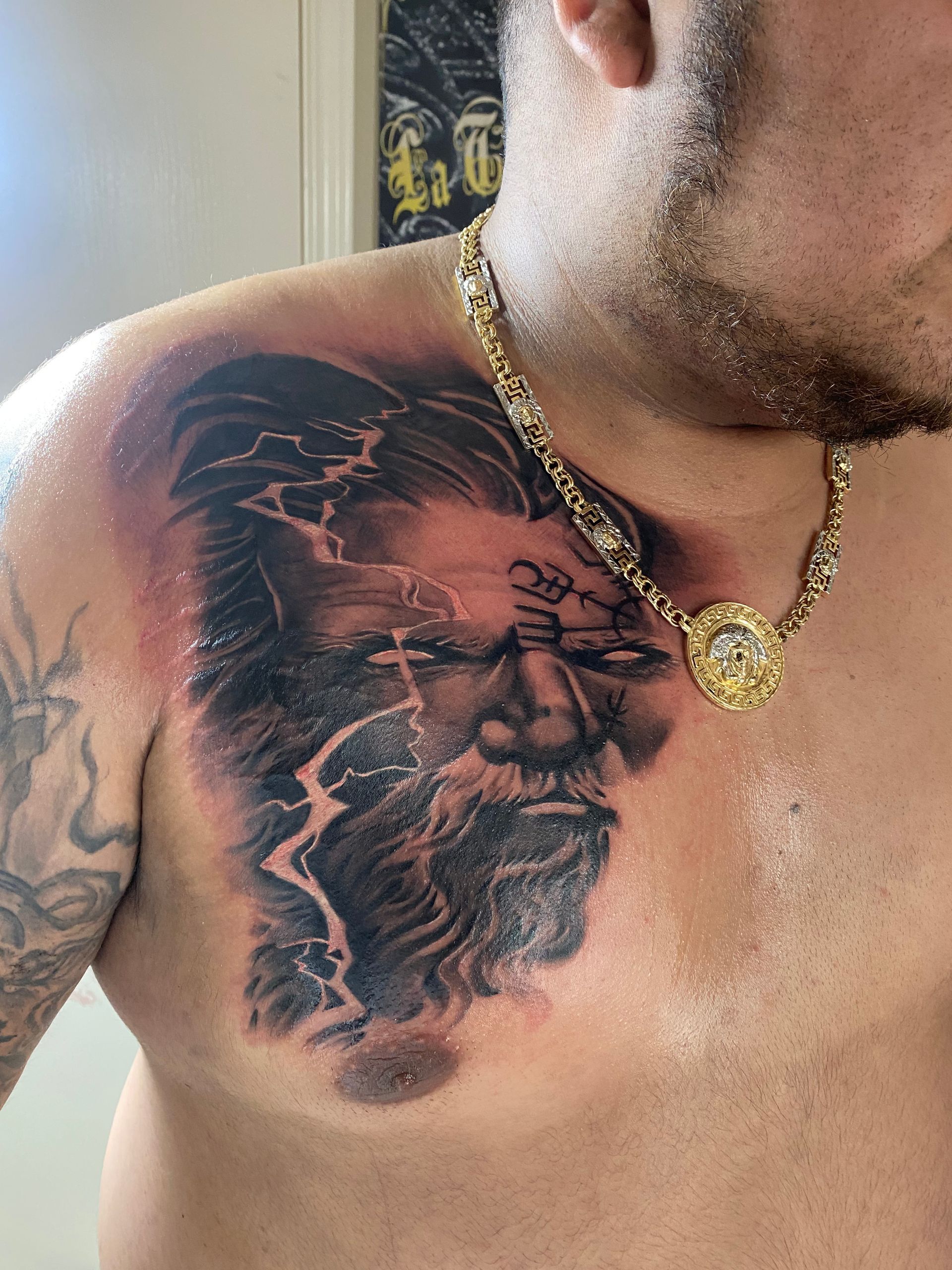 Man with chest tattoo of Zeus, lightning, and dark colors, wearing a gold chain.