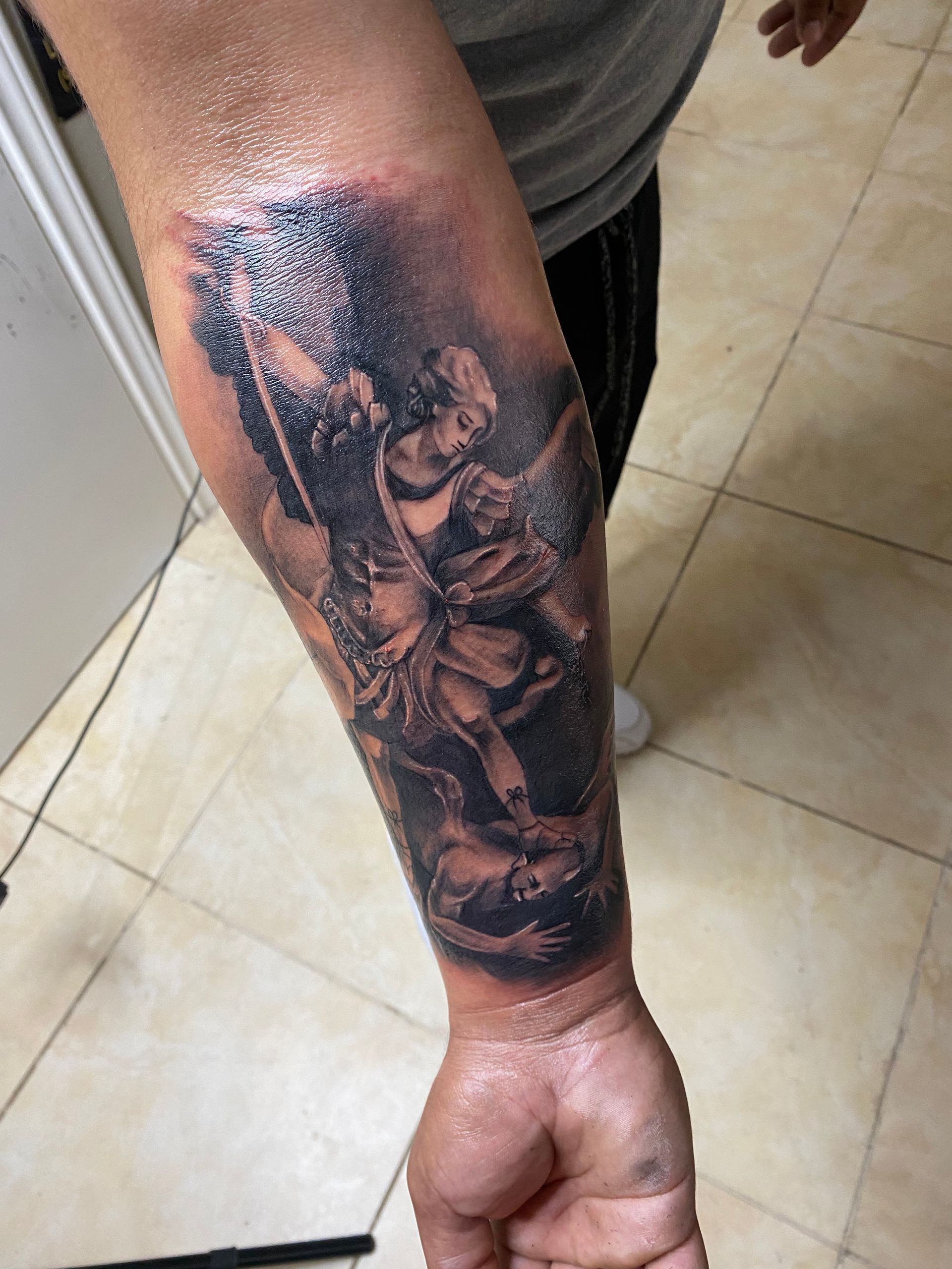 Tattoo of Saint Michael defeating the devil on a forearm in black and gray.